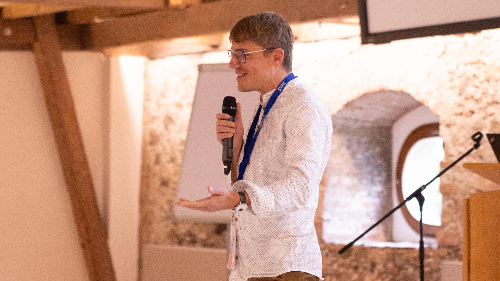 A person wearing glasses and a white shirt speaks into a microphone while standing indoors near a window and wooden beams, gesturing with one hand. A presentation screen and microphone stand are visible in the background.