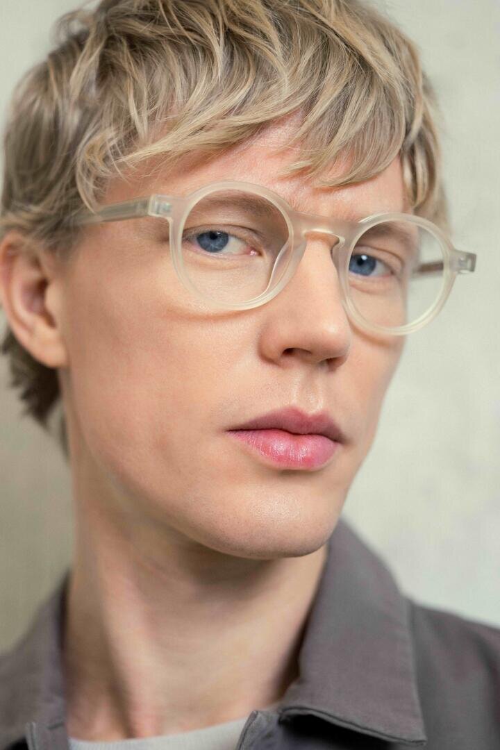 Eine Person mit kurzem, hellblondem Haar und einer klaren, runden Brille blickt leicht an der Kamera vorbei. Sie hat helle Haut, blaue Augen und trägt ein graues Hemd mit Kragen vor einem neutralen Hintergrund.