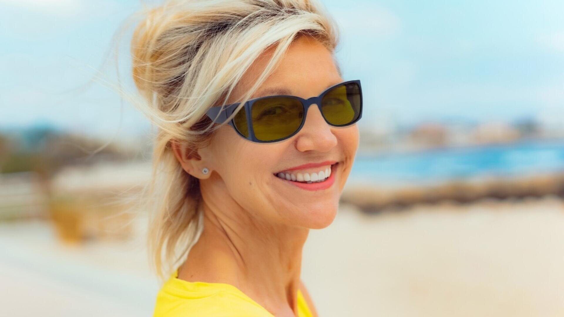 Smiling woman with blonde hair wearing dark sunglasses and a yellow shirt stands outdoors near a beach on a sunny day. Her hair is windblown, and the background is bright and slightly blurred.