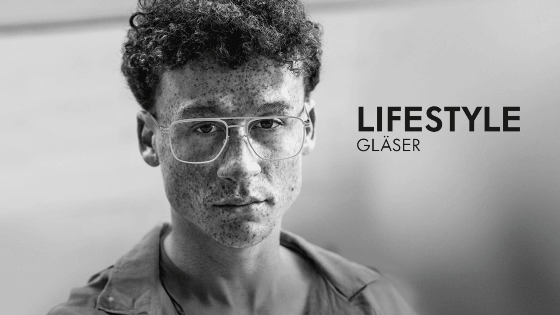 Schwarz-Weiß-Foto einer jungen Person mit lockigem Haar, Sommersprossen und großer Brille, die in die Kamera blickt. Rechts ist fettgedruckt der Schriftzug „LIFESTYLE GLÄSER“ zu sehen.