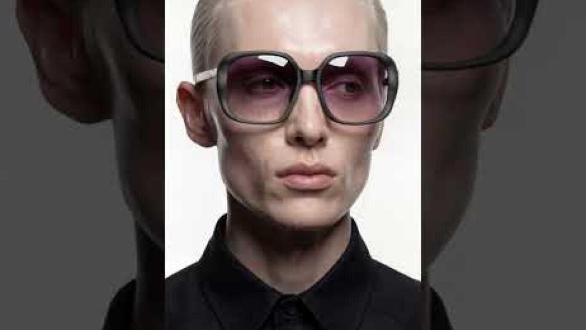 Eine Person mit kurzen blonden Haaren trägt eine große, dunkle Sonnenbrille und ein schwarzes Hemd mit Kragen und blickt leicht zur Seite vor einem schlichten, hellen Hintergrund.