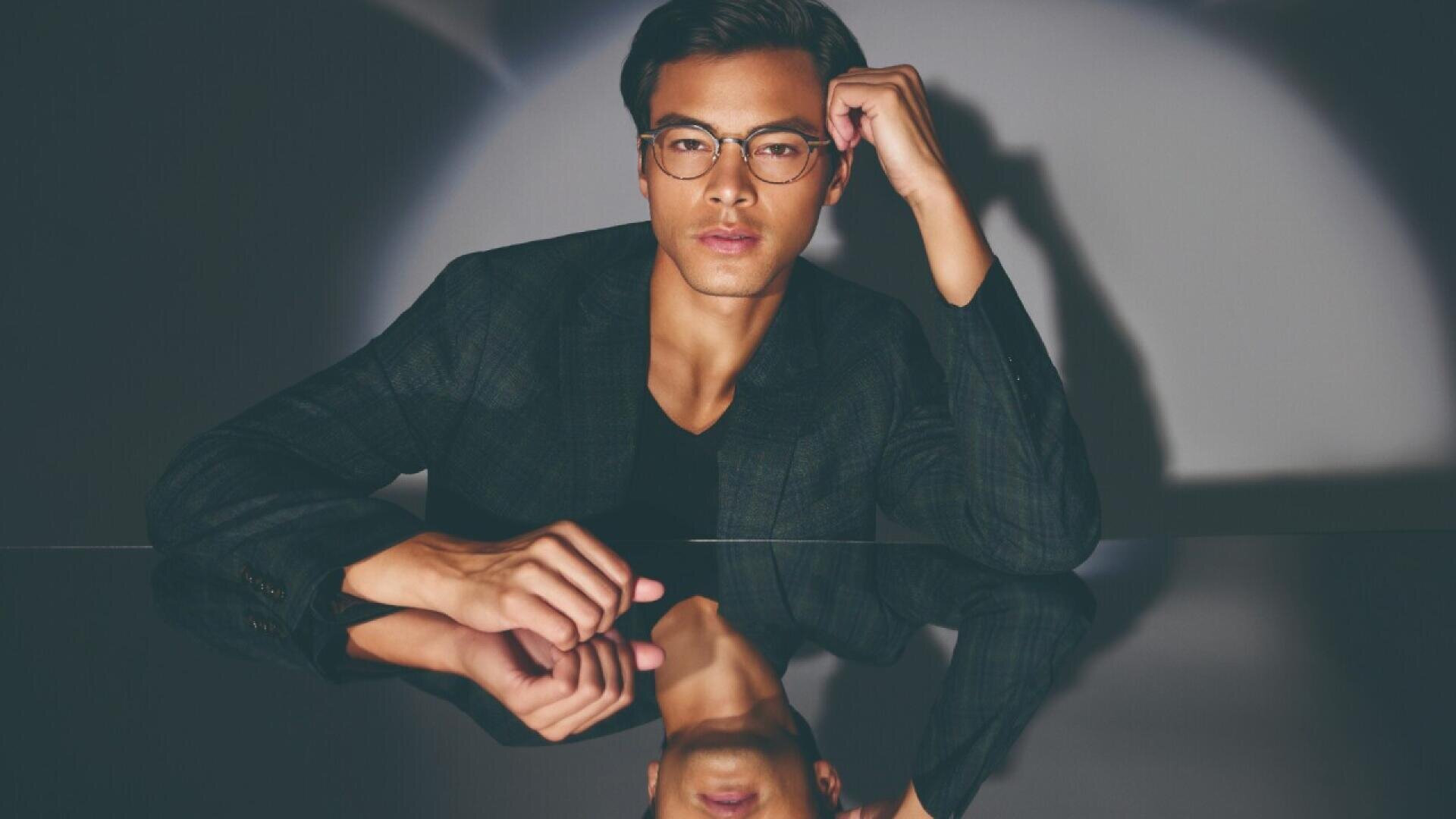 A man wearing glasses and a dark blazer sits at a reflective surface, resting one arm on the table and the other hand near his face, with his reflection visible below. The background is softly lit.