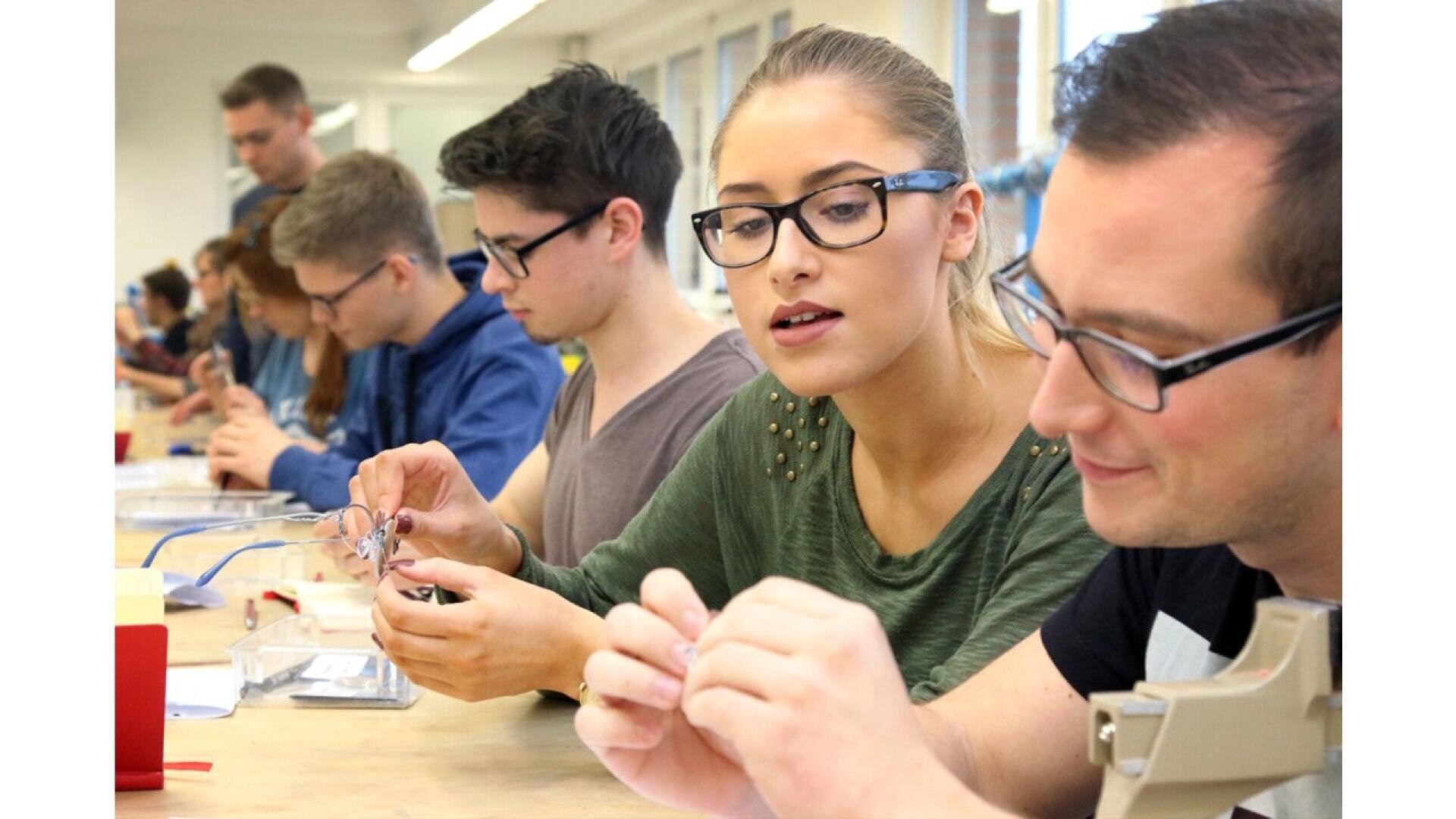 A group of people, mostly young adults, sit at a long table working on small projects with their hands. They appear focused and are wearing glasses, suggesting a classroom or workshop setting.