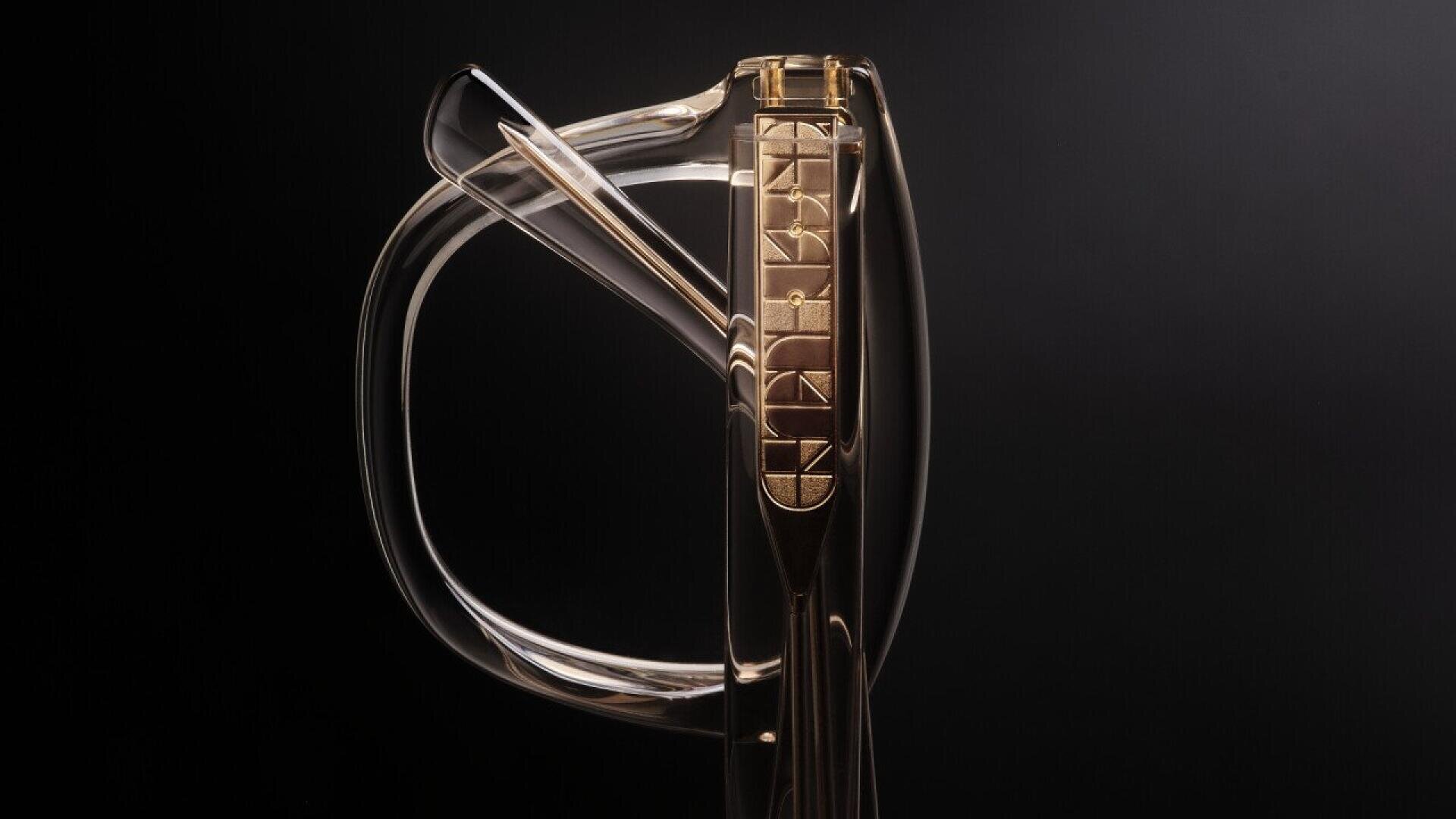 A close-up of a transparent glass sword with a golden hilt featuring geometric engravings, set against a dark background.