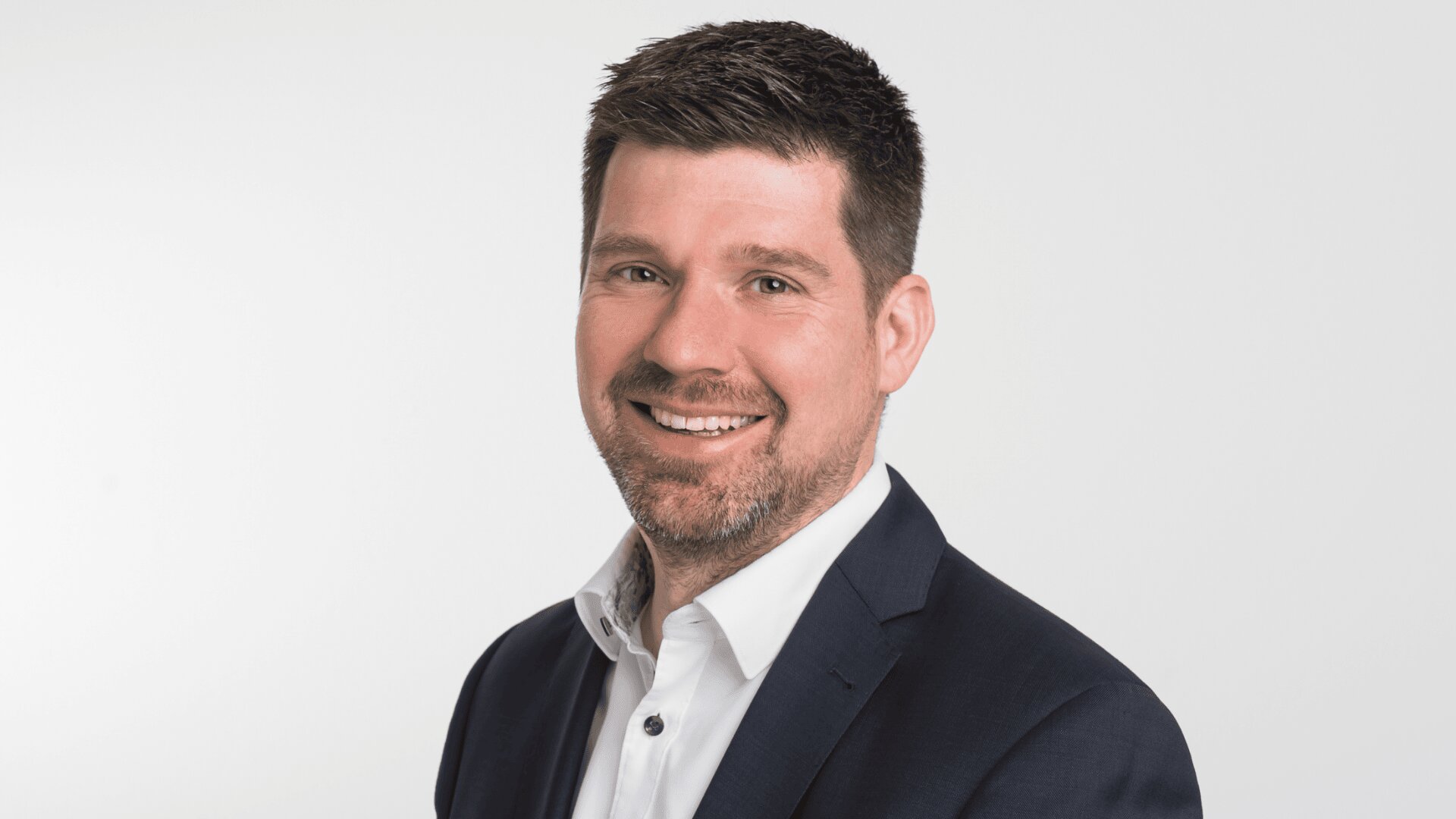 Ein Mann mit kurzen dunklen Haaren und gestutztem Bart, der ein weißes Hemd und einen dunklen Blazer trägt und vor einem schlichten weißen Hintergrund lächelt.