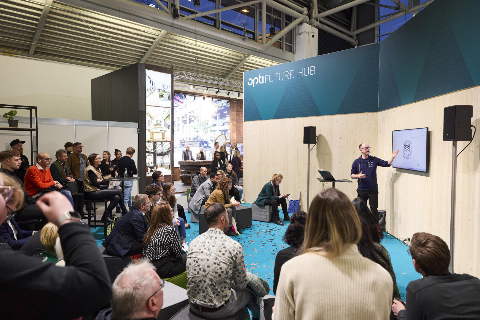 A speaker speaks to an attentive audience in a modern event room labeled FUTURE HUB with a cartoon graphic on the screen. People sit and stand, listen and take notes.