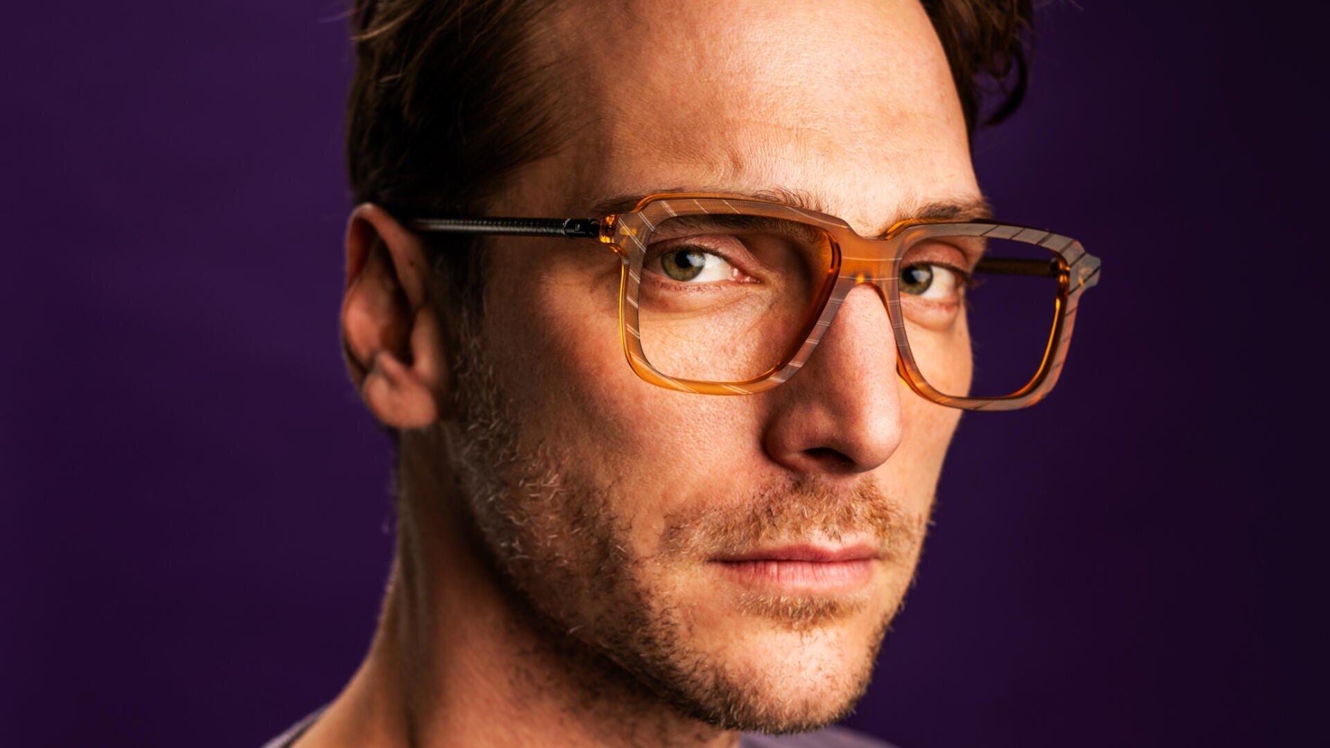A man with short brown hair and light facial stubble wears large, square, amber glasses and looks seriously at the camera against a solid purple background.
