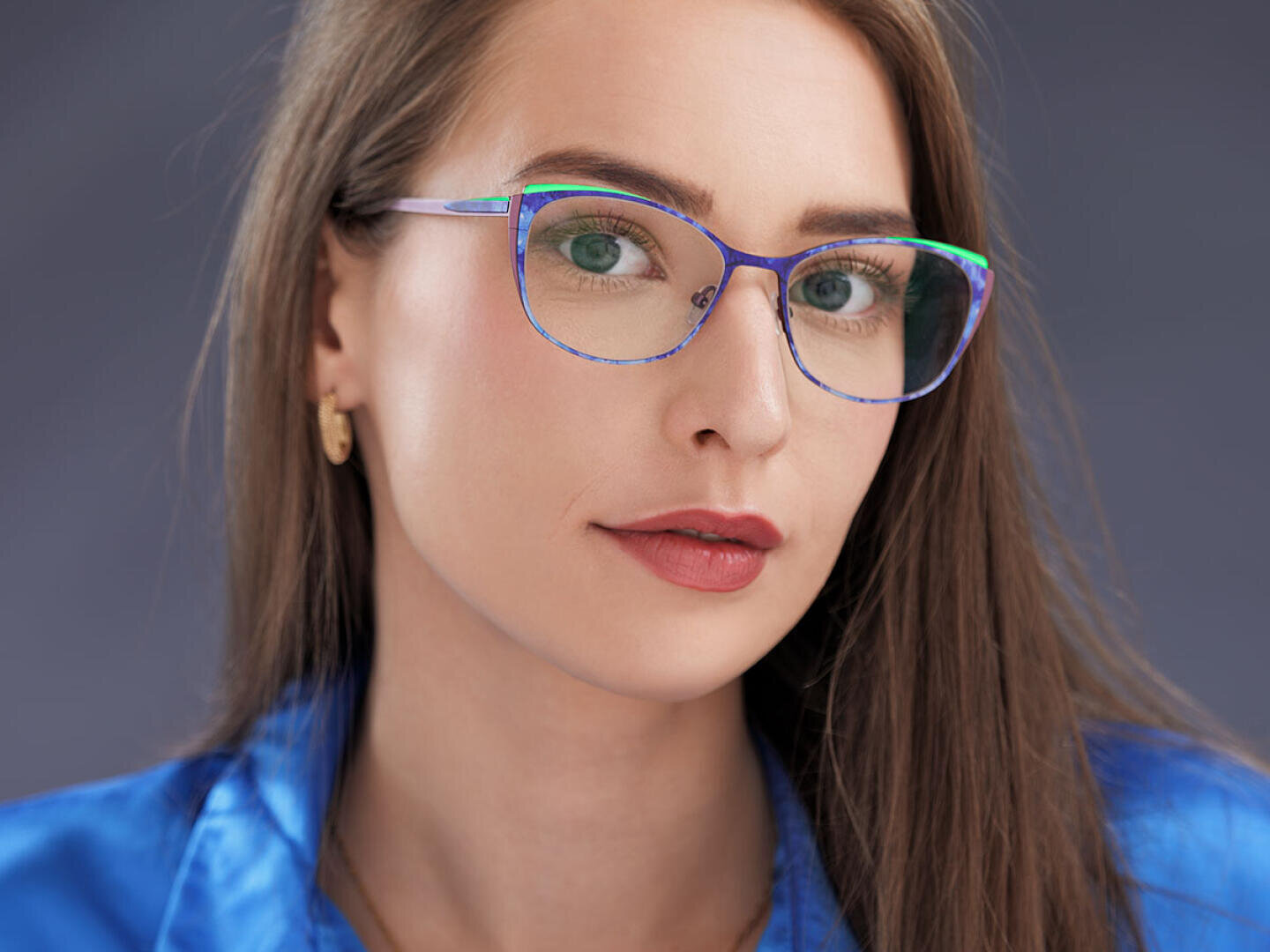 A woman with long brown hair and green eyes wears colorful glasses, gold earrings and a blue satin shirt. She looks directly into the camera with a neutral expression against a plain dark background.