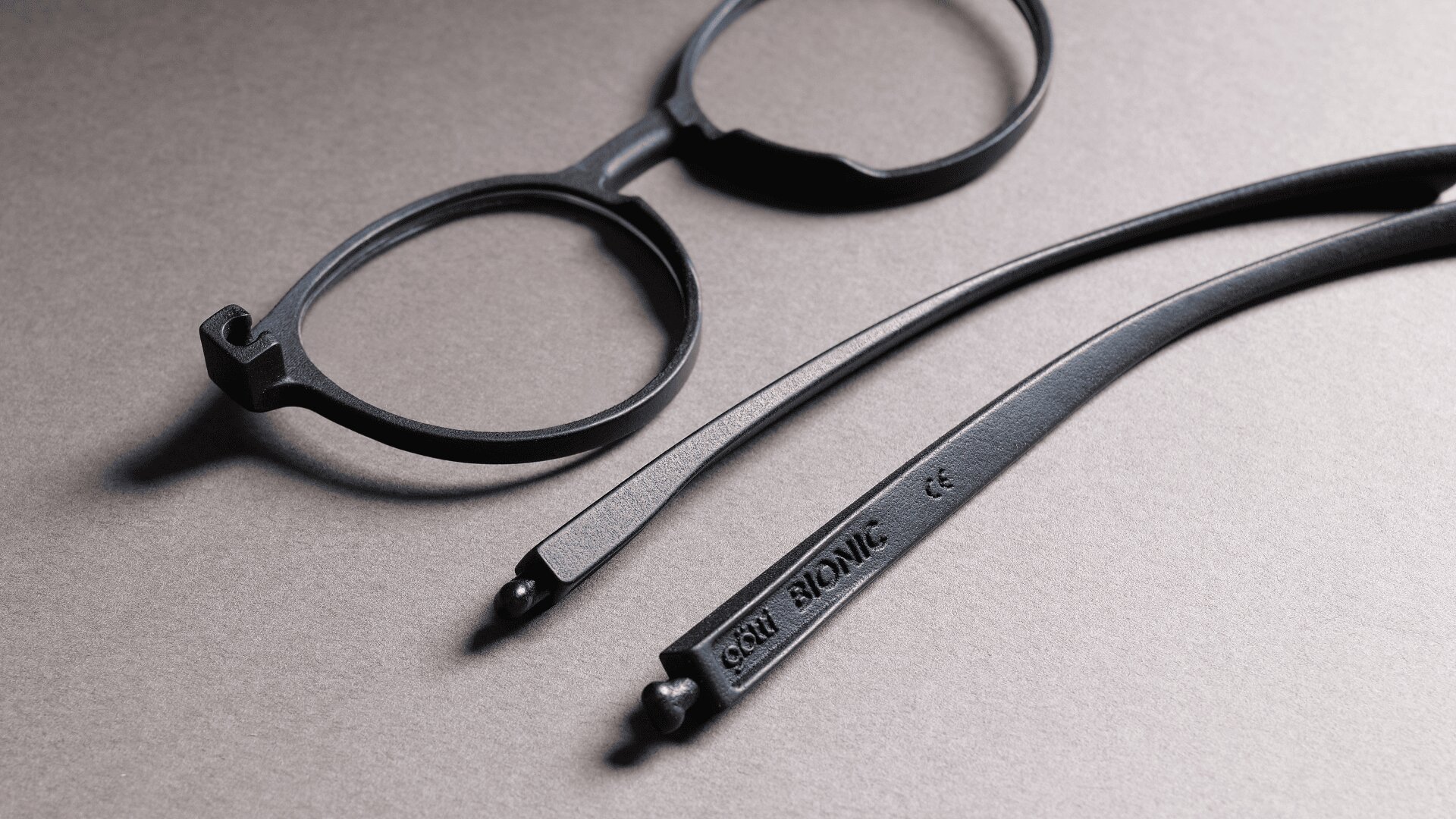 Ein schwarzes Brillengestell mit abgenommenen Bügeln, das auf einer grauen Fläche liegt. Einer der Bügel ist mit der Aufschrift BIONIC und zusätzlichen Markierungen und Zahlen versehen.