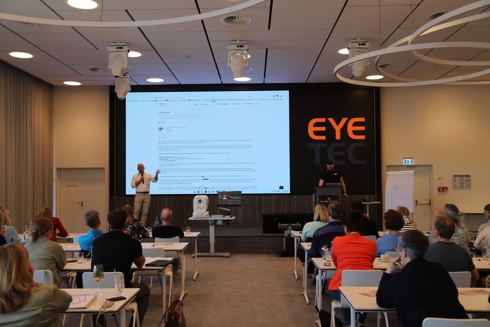Ein Redner steht auf der Bühne und hält einen Vortrag vor einem Publikum, das an Tischen in einem Konferenzraum sitzt. Auf einem großen Bildschirm wird eine Webseite angezeigt, und eine weitere Person steht an einem Podium. An der Wand dahinter ist der Text EYE TEC zu lesen.