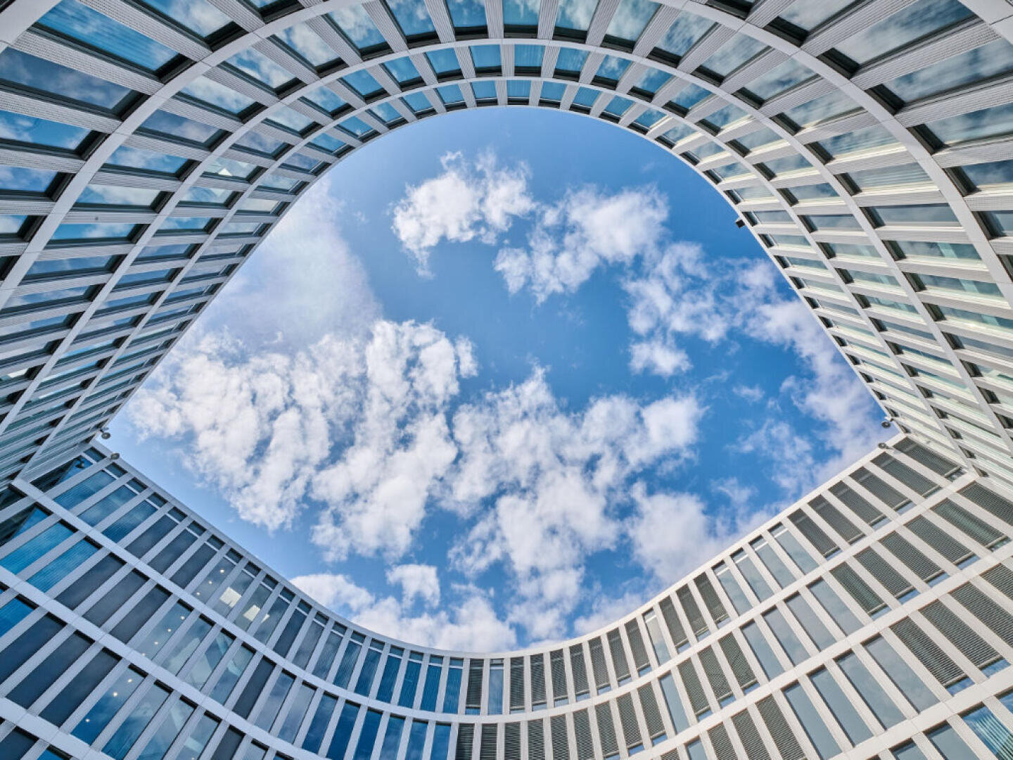 Ein modernes Gebäude mit geschwungenen Glasfenstern umgibt einen offenen ovalen Raum, über dem ein blauer Himmel mit vereinzelten Wolken zu sehen ist. Das Foto wurde vom Boden aus aufgenommen, wobei der Blick gerade nach oben gerichtet ist.