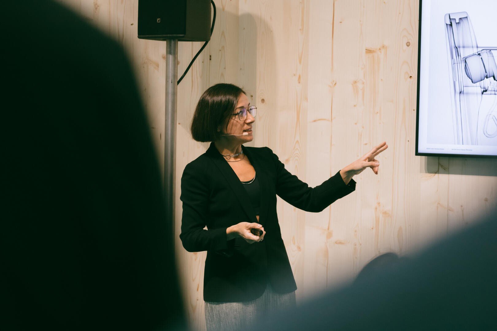 A woman in a black blazer and glasses gives a lecture and points to a screen on which a technical drawing can be seen, with a light wood-paneled wall in the background.