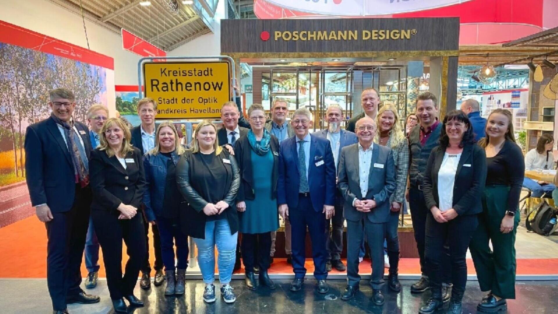 A group of people in business attire pose and smile in front of a Poschmann Design booth at an indoor event. A yellow road sign reads “Kreisstadt Rathenow Stadt der Optik Landkreis Havelland.”.