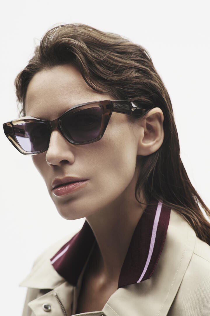 A woman with wavy brown hair wears dark cat-eye sunglasses and a beige jacket with a maroon and white striped collar, looking confidently at the camera against a plain white background.
