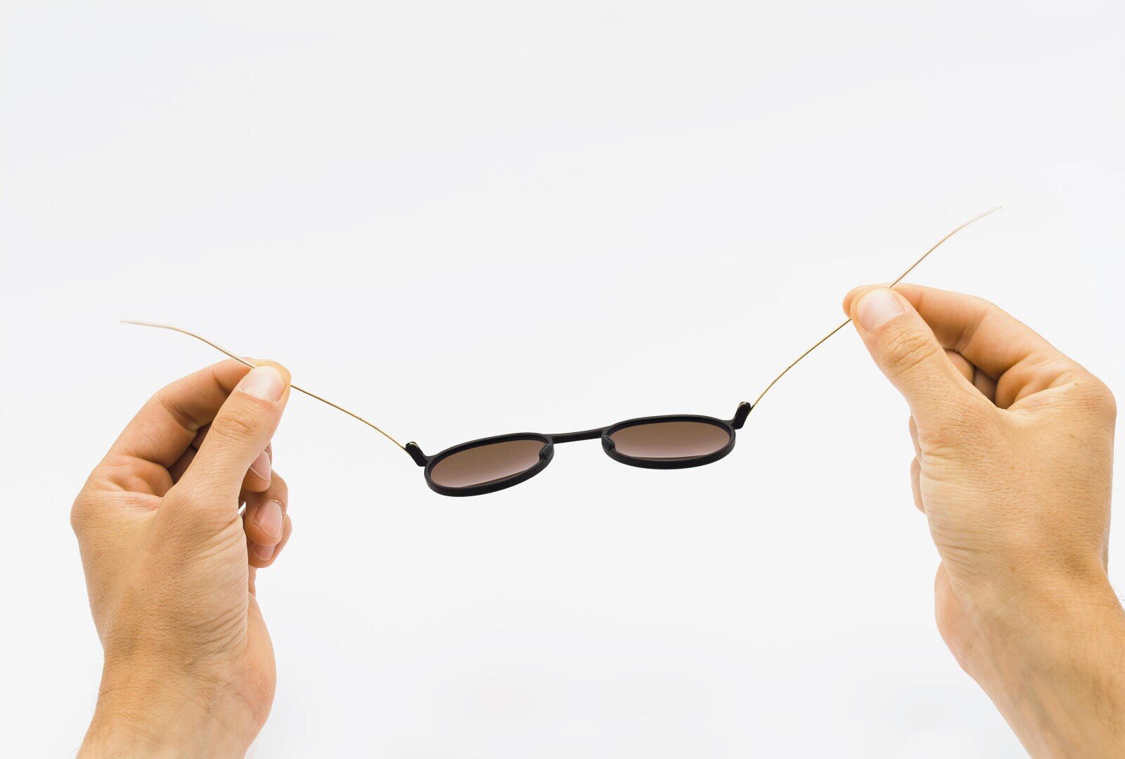 Zwei Hände halten eine Sonnenbrille mit dünnen, gebogenen Drahtbügeln vor einem weißen Hintergrund.