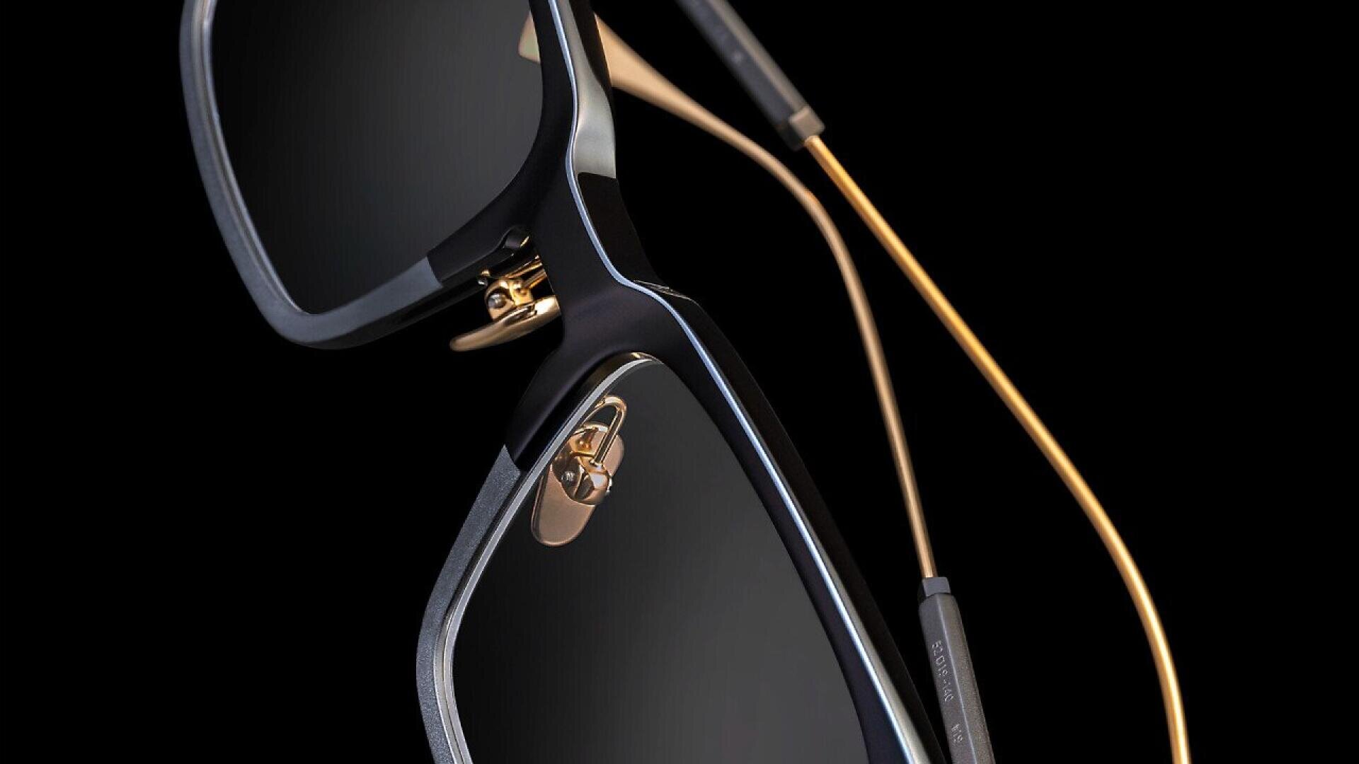 Close-up of a pair of stylish sunglasses with dark lenses, thin gold-colored arms, and black frames, set against a black background.