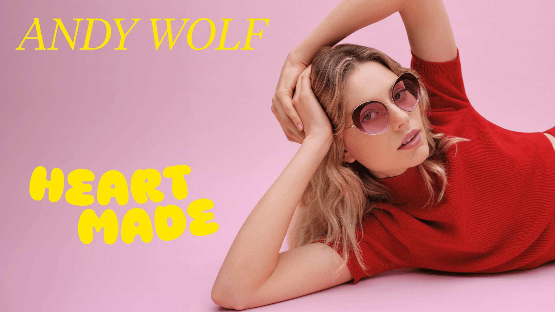 Eine Frau in einem roten Oberteil posiert auf einer rosa Fläche und trägt eine runde rosa Sonnenbrille. Oben links steht in kräftigem Gelb ANDY WOLF und links in verspielter Schrift HEART MADE.