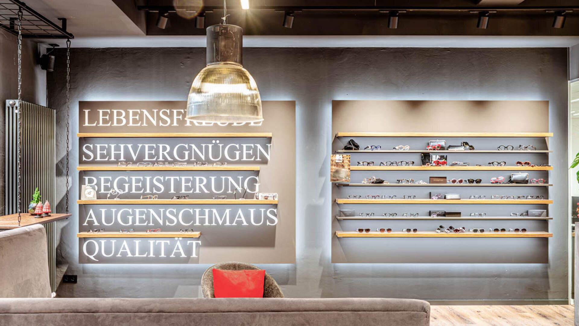 Modernes Interieur eines Brillengeschäfts mit beleuchteten deutschen Schriftzügen an der linken Wand und zwei Regalen mit verschiedenen Brillen und Sonnenbrillen an der rechten Wand. Eine Hängelampe und ein graues Sofa mit einem roten Kissen sind ebenfalls zu sehen.