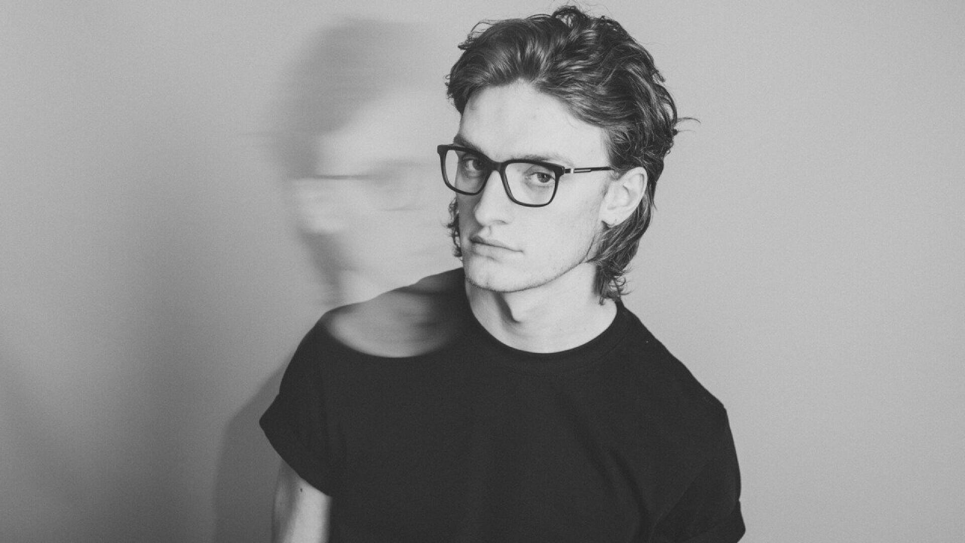 A young man wearing glasses and a black t-shirt stands against a plain background with a blurred double-exposure effect showing his face in profile to the left.