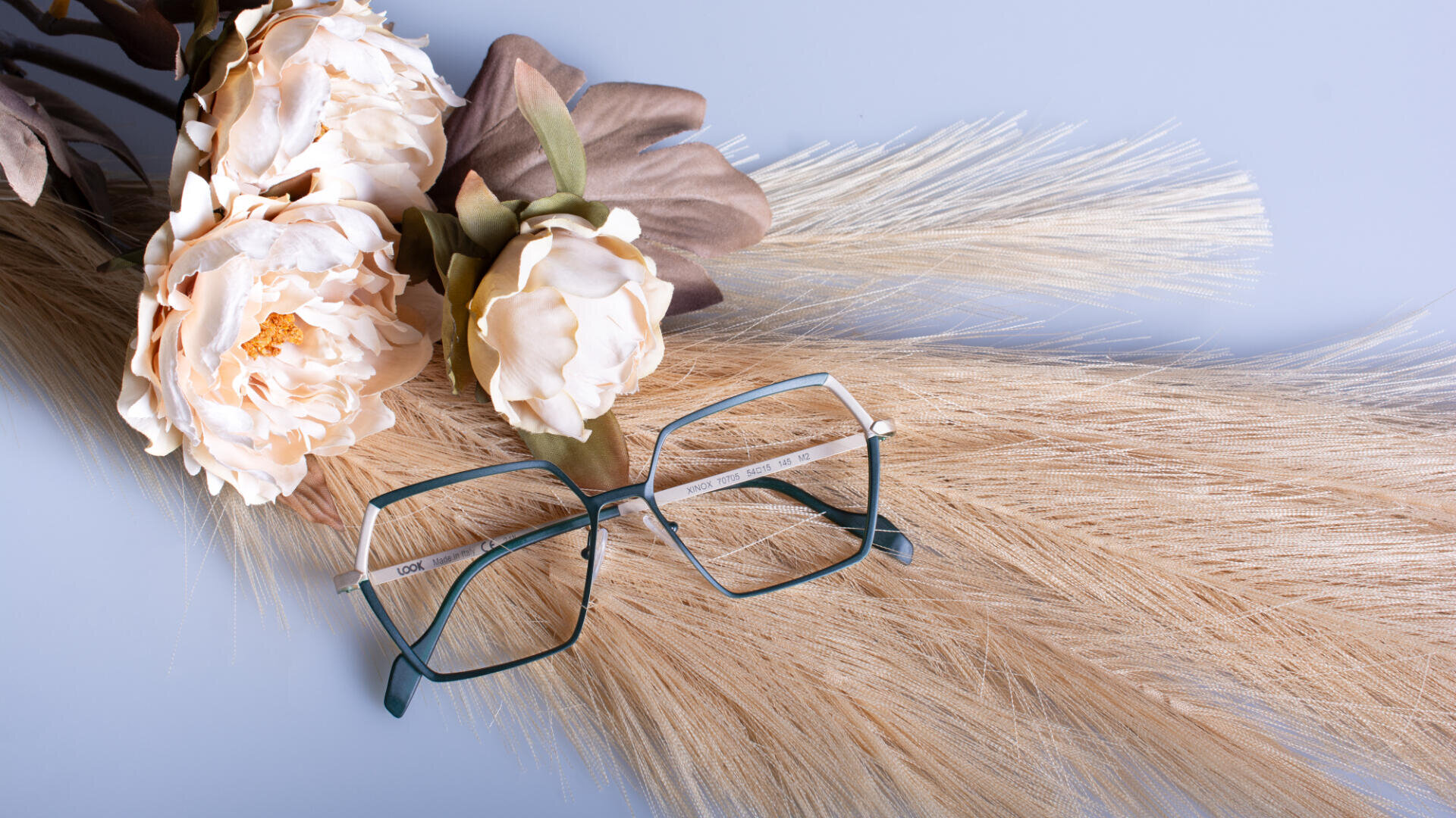 A pair of rectangular eyeglasses with dark green frames rests on dried wheat stalks, next to artificial cream and brown flowers, all set against a soft light blue background.