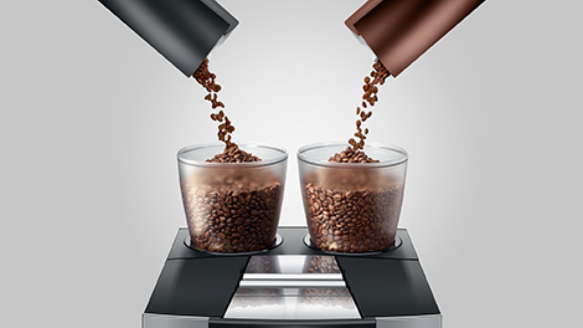 Zwei transparente, mit Kaffeebohnen gefüllte Gläser stehen unter zwei Metallzylindern, in die jeweils weitere Kaffeebohnen gegossen werden. Das Ganze steht vor einem schlichten, hellen Hintergrund.