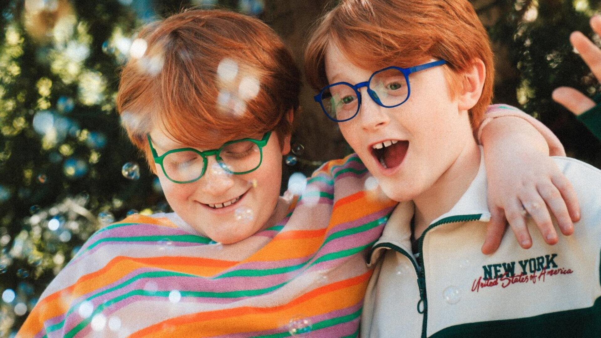 Two smiling children with red hair and glasses stand close together outdoors, arm in arm, surrounded by sunlight and bubbles. One wears a striped sweater, the other a white jacket with New York text.