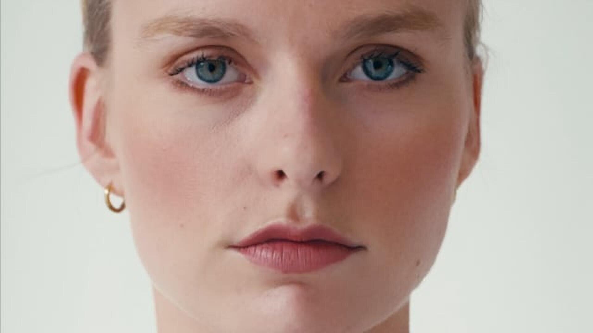 A close-up of a person with fair skin, blue eyes, and light pink lips, looking directly at the camera. They have minimal makeup and are wearing a small gold hoop earring. The background is plain and light-colored.