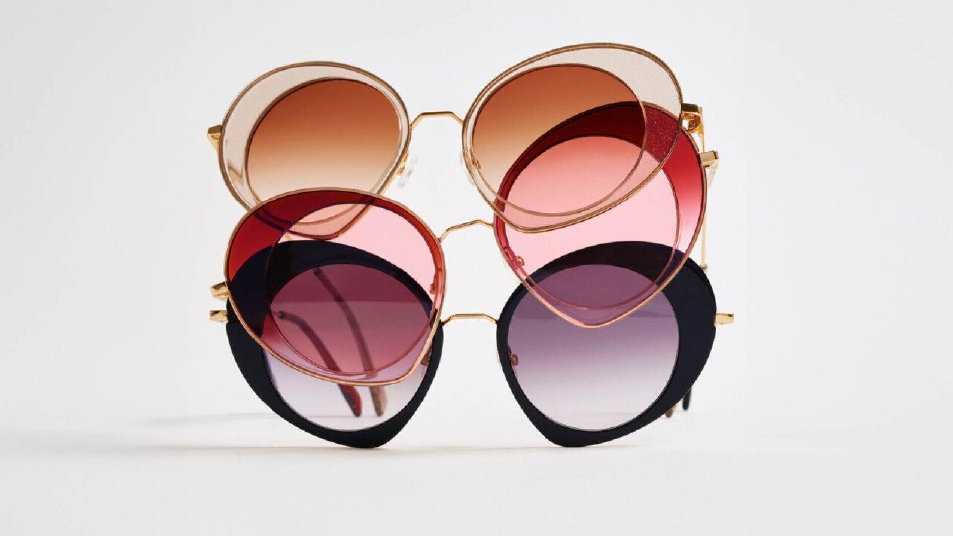 Three pairs of cat-eye sunglasses with gold frames, stacked vertically. The top has brown lenses, the middle has pink lenses, and the bottom has dark purple lenses with black rims, all on a white background.