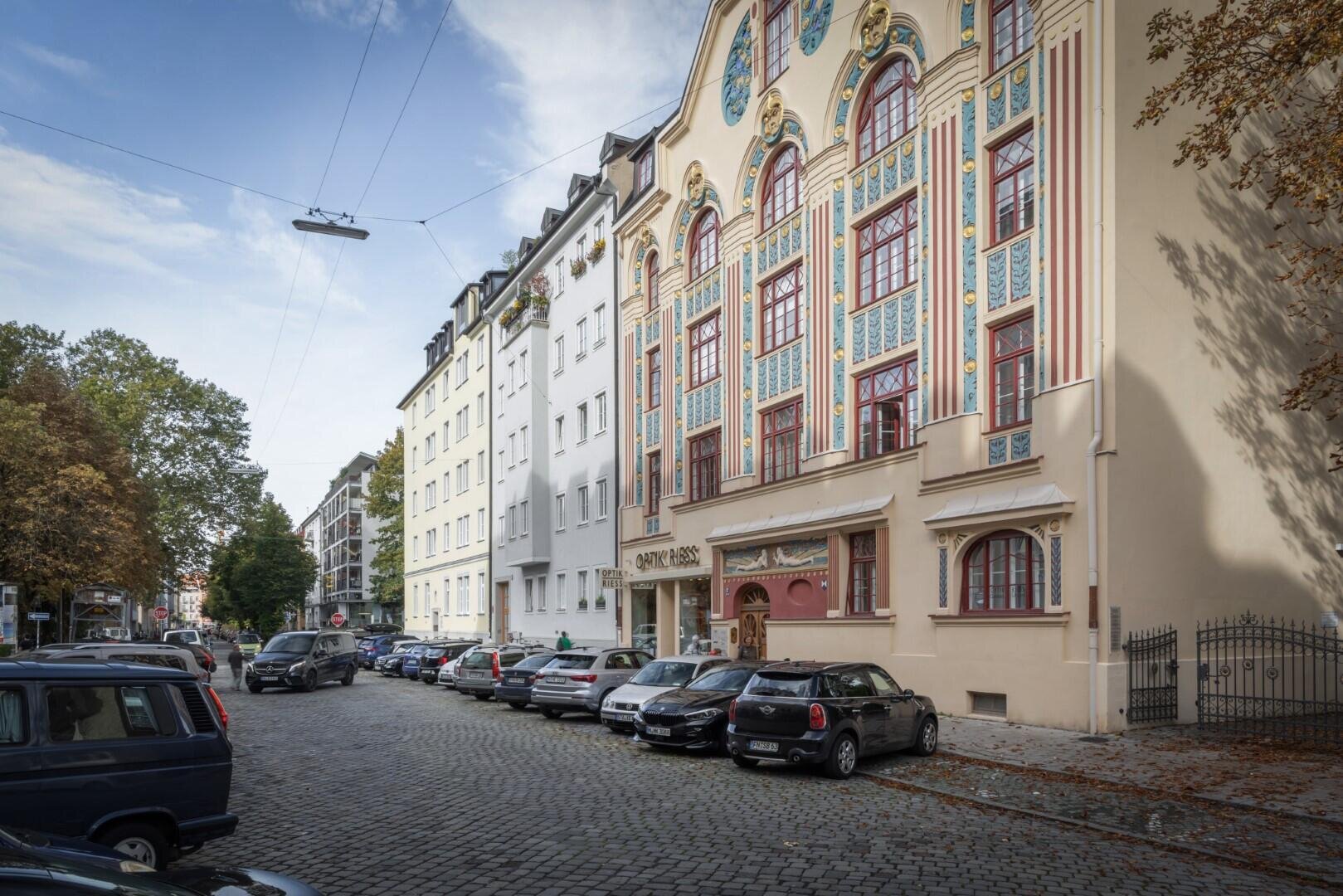 Eine Straße mit parkenden Autos neben hohen Gebäuden, darunter ein verziertes beiges Gebäude mit farbenfrohen, dekorativen Fenstern und detailreichen architektonischen Details. Bäume säumen die Straße, der Himmel ist teilweise bewölkt.