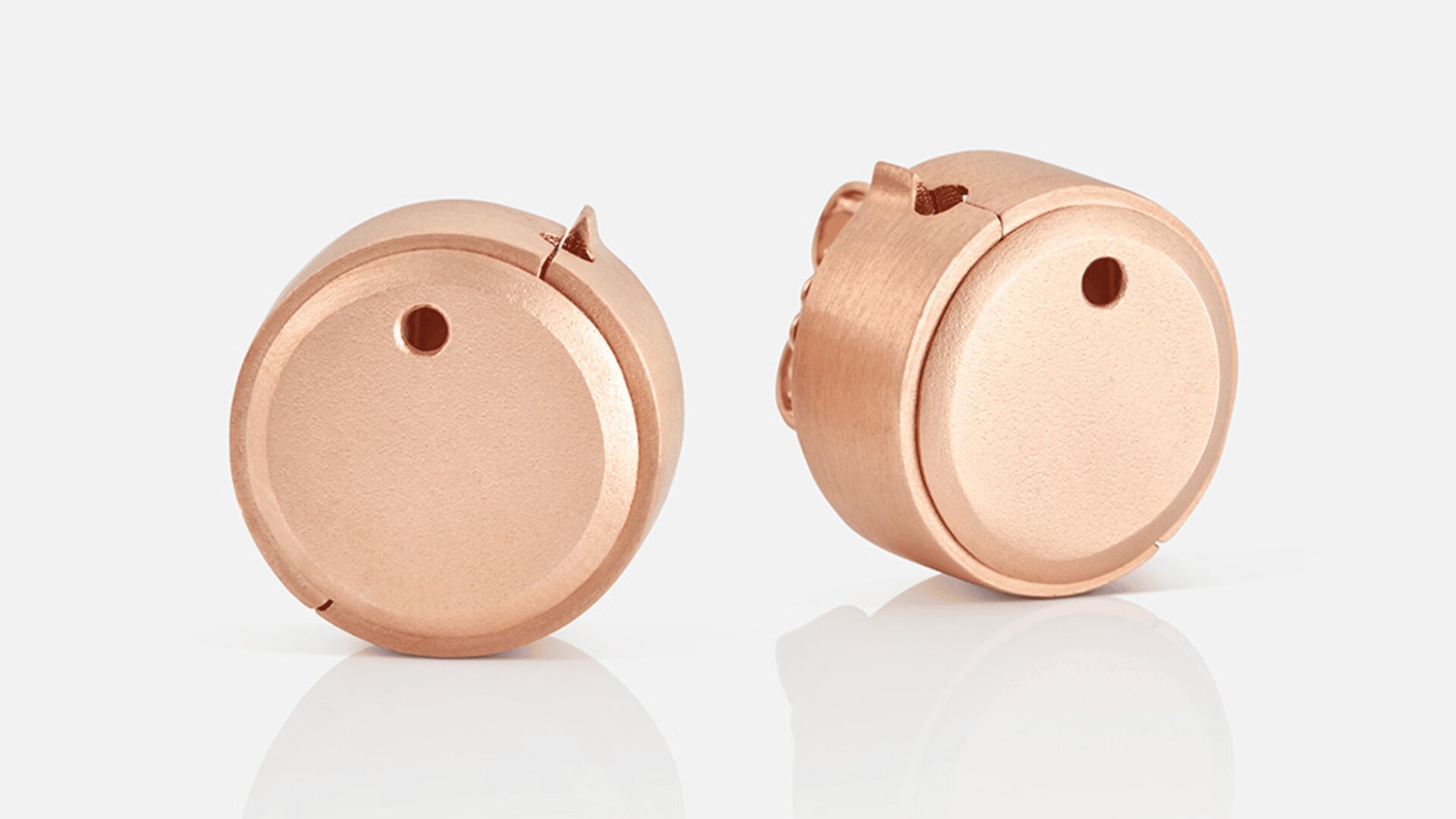 Zwei kleine, runde, roségoldfarbene Objekte mit glatten Oberflächen und kleinen Löchern auf weißem Hintergrund. Ein Objekt steht aufrecht, das andere ist leicht geneigt, sodass an der Seite strukturierte Details sichtbar sind.