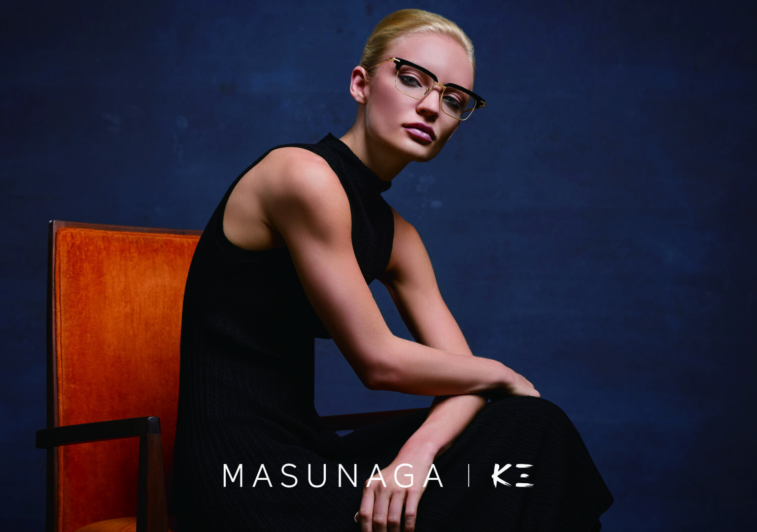Eine Frau mit Brille und einem ärmellosen schwarzen Kleid sitzt auf einem orangefarbenen Stuhl vor einem dunkelblauen Hintergrund. Der Schriftzug MASUNAGA | KE ist am unteren Rand zu sehen.