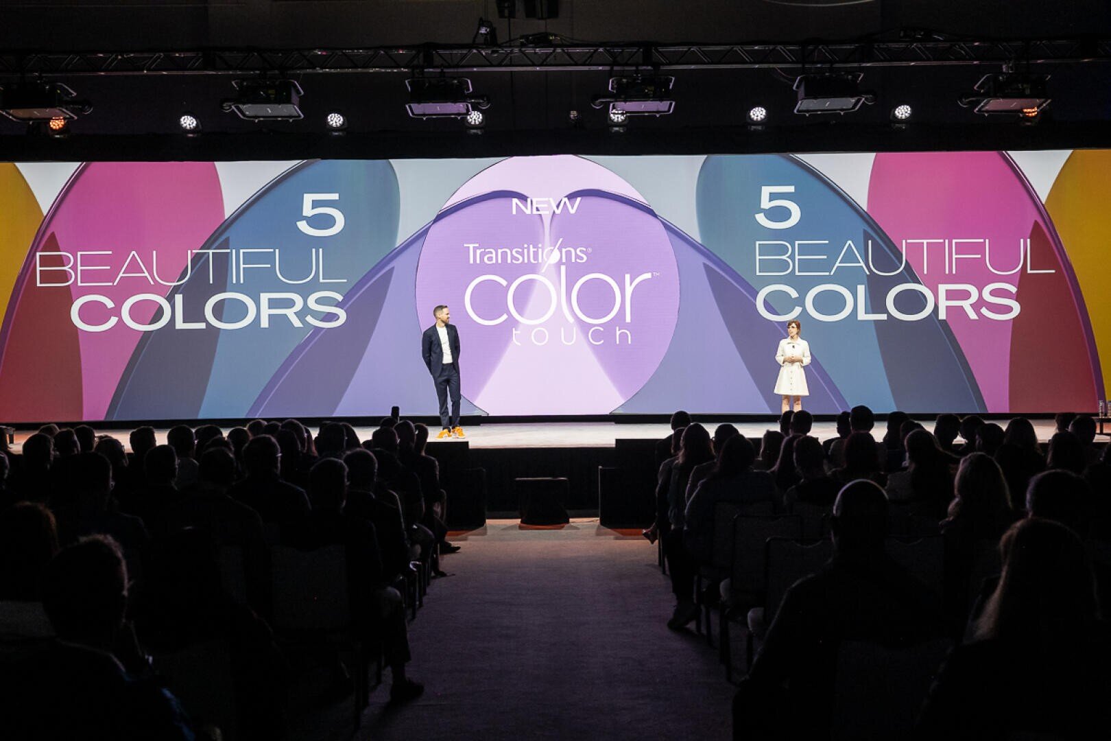 Zwei Personen stehen auf einer Bühne und stellen sich dem Publikum vor. Hinter ihnen wird auf einem großen Bildschirm "NEW Transitions Color Touch" mit der Aufschrift "5 Beautiful Colors" auf beiden Seiten in bunten Texten und Grafiken angezeigt.