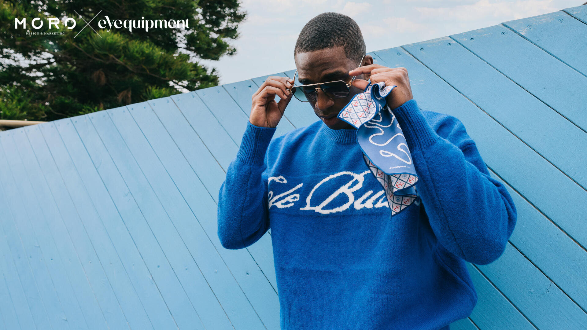 Ein Mann in einem leuchtend blauen Pullover mit weißer Schrift rückt seine Sonnenbrille zurecht, während er ein gemustertes Tuch hält und im Freien vor einer schrägen, hellblauen Holzwand steht.