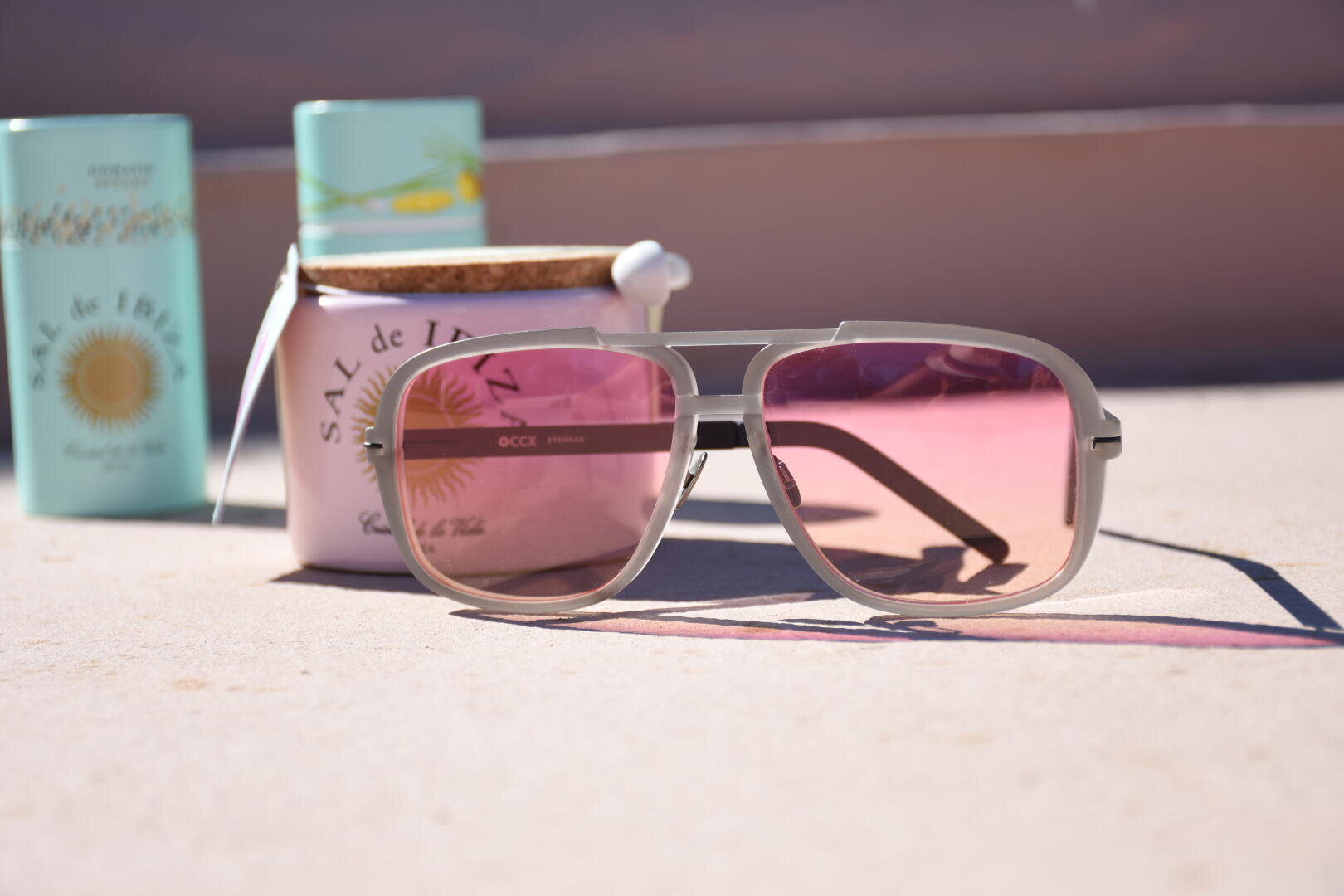 Eine hellgraue Sonnenbrille mit rosa getönten Gläsern liegt auf einer hellen Fläche, im Hintergrund stehen pastellfarbene Behälter mit der Aufschrift Sal de Ibiza und ein Glas mit Korkdeckel.