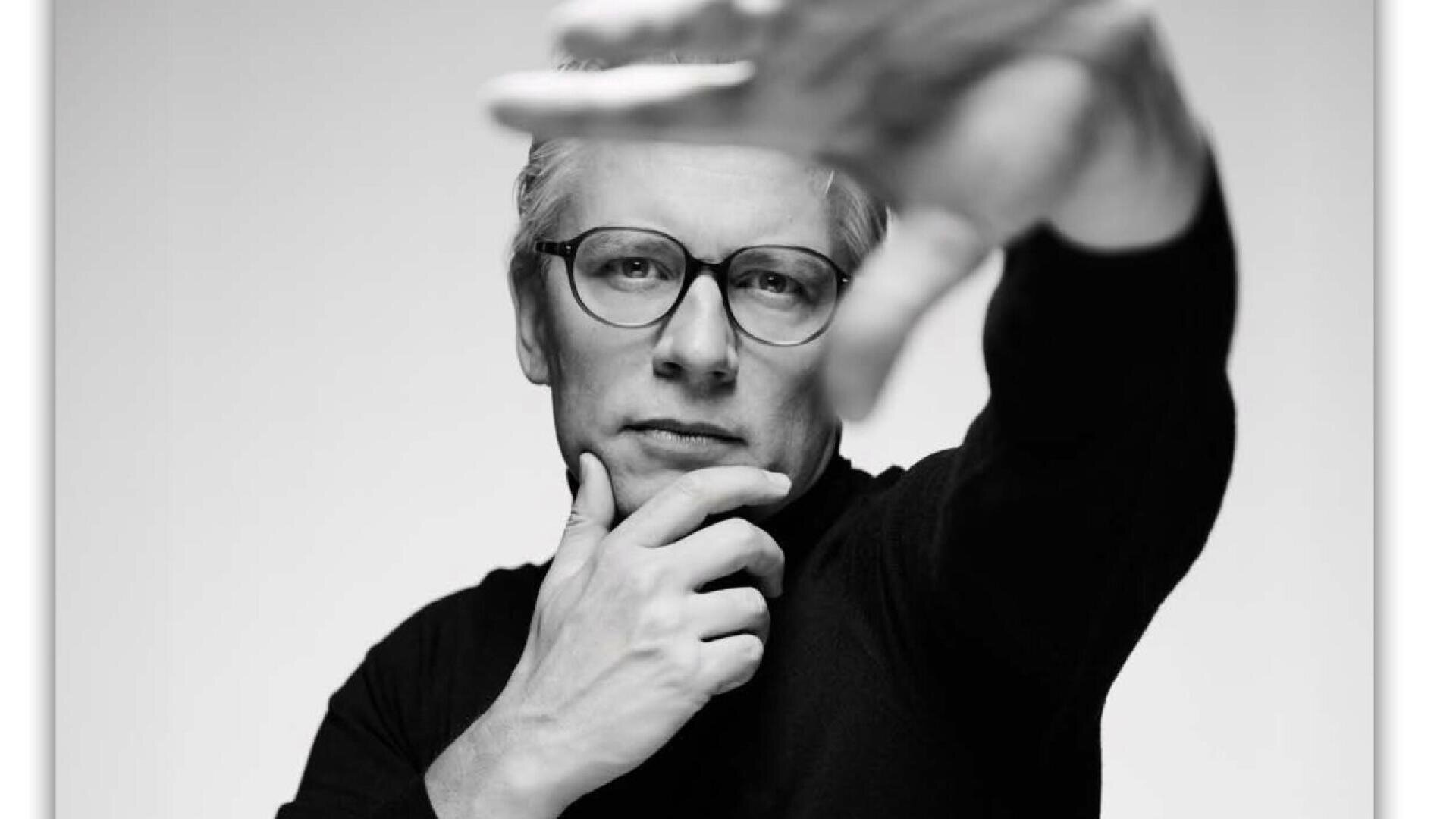 A person wearing glasses looks thoughtfully at the camera, resting one hand on their chin while extending the other hand forward, partially obscuring their face. The image is in black and white.