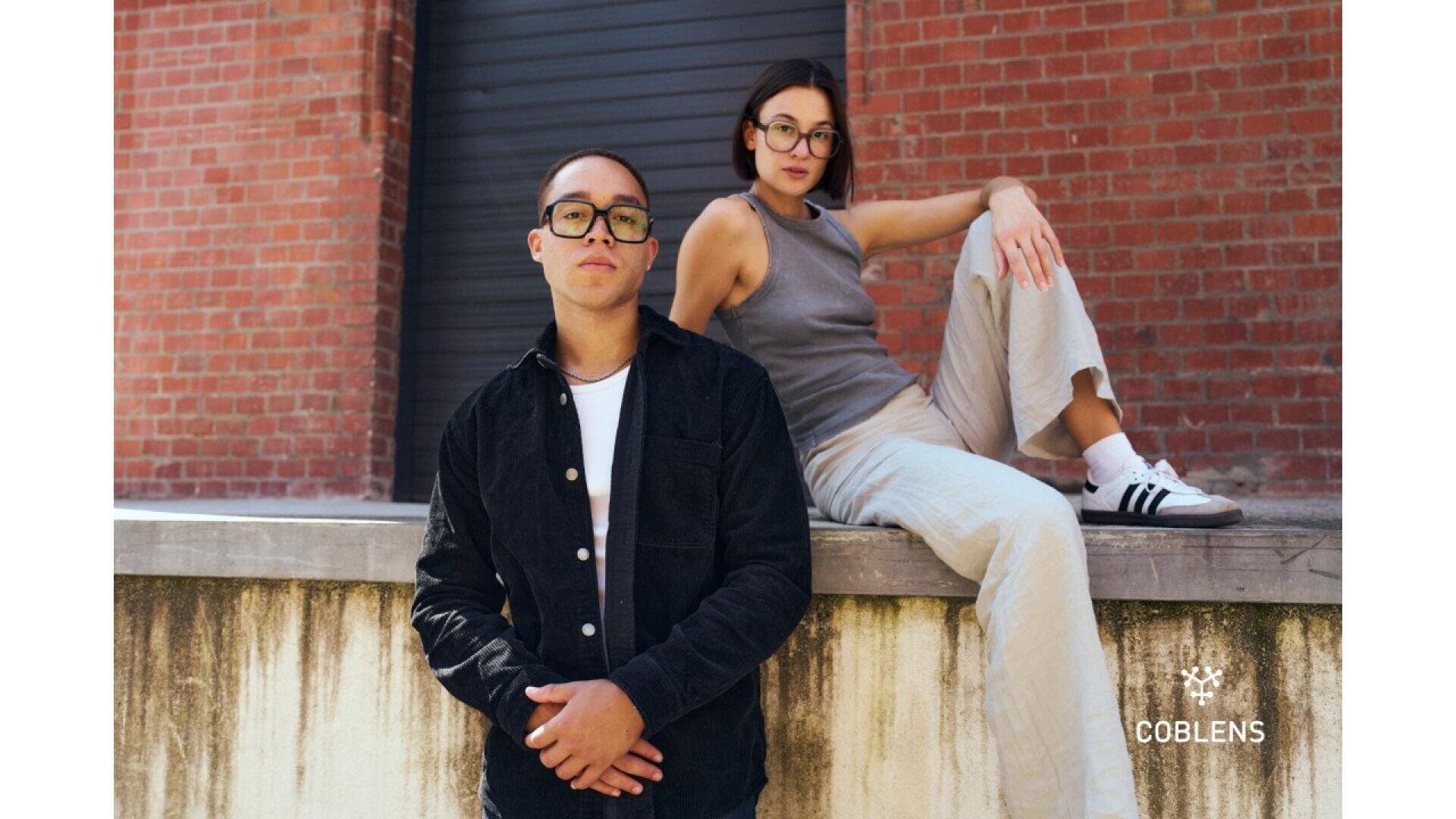 Two people wearing glasses pose outdoors against a brick wall; one stands in a black jacket while the other sits on the ledge in a gray top and beige pants. The logo “COBLENS” appears in the corner.