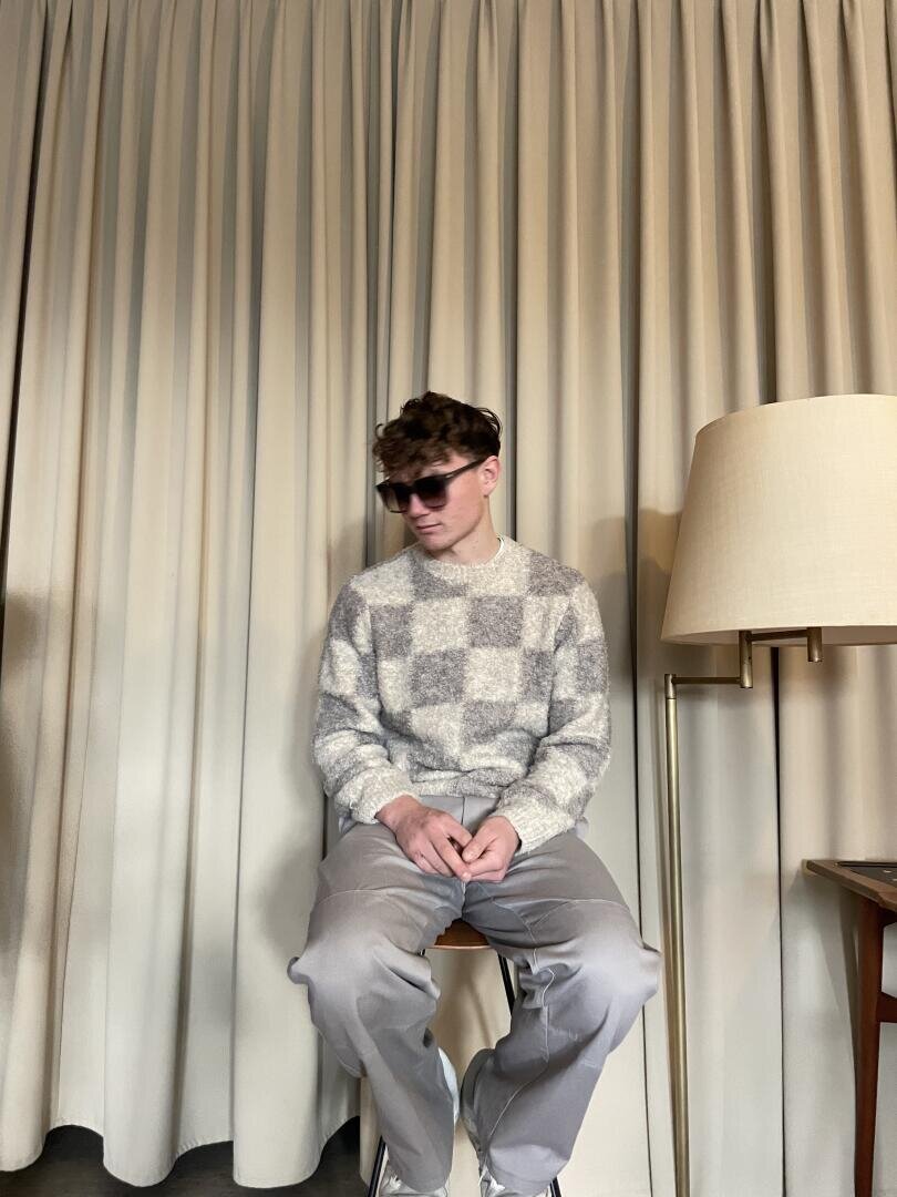 Ein junger Mann, der eine Sonnenbrille, einen grau-weiß karierten Pullover und eine hellgraue Hose trägt, sitzt auf einem Hocker vor beigen Vorhängen neben einer Stehlampe.