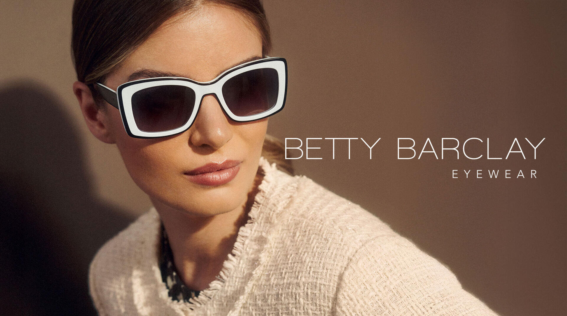 Eine Frau mit einer großen, quadratischen, schwarz-weißen Sonnenbrille und einer strukturierten cremefarbenen Jacke posiert vor einem braunen Hintergrund. Der Text lautet BETTY BARCLAY EYEWEAR.