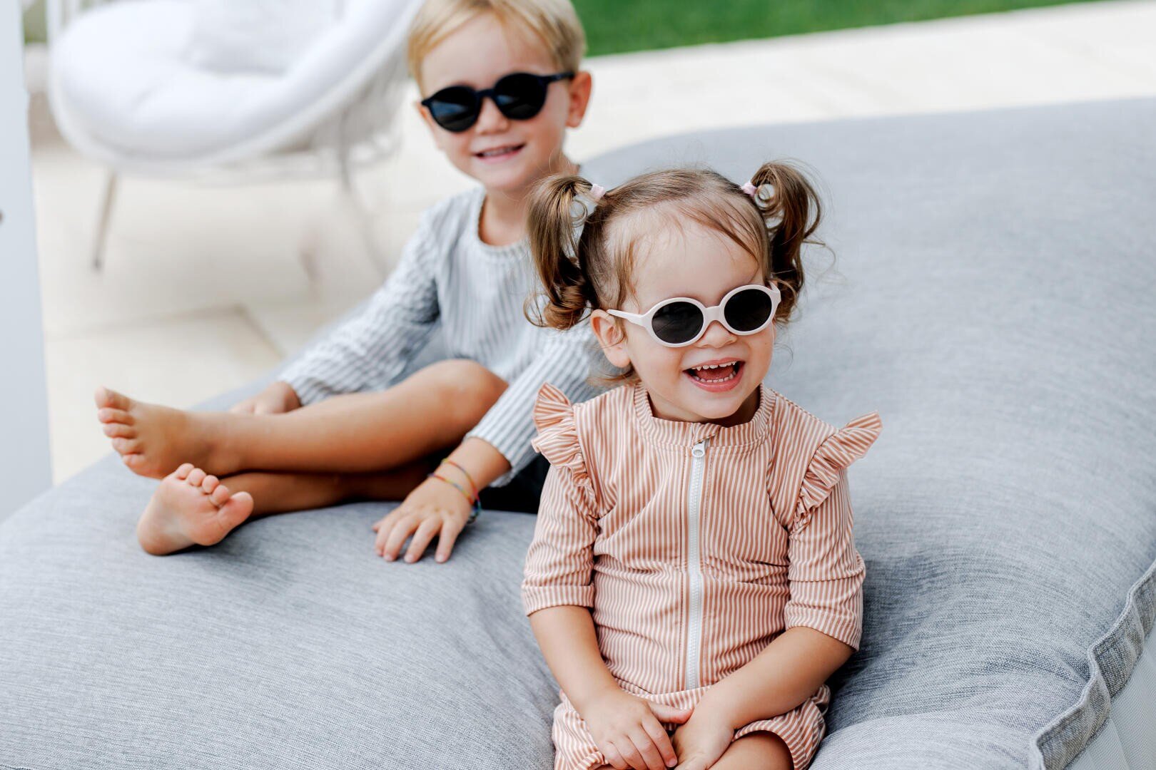 Two small children with sunglasses and striped clothes are sitting on a large gray cushion outside. They are smiling and enjoying a sunny day, with one child in the foreground and the other behind them.