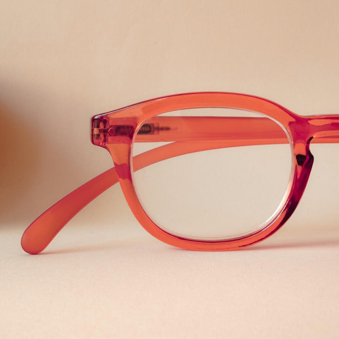 Eine Nahaufnahme eines roten Brillengestells mit klaren Gläsern, das auf einer hellbeigen Fläche und einem hellbeigen Hintergrund ruht.