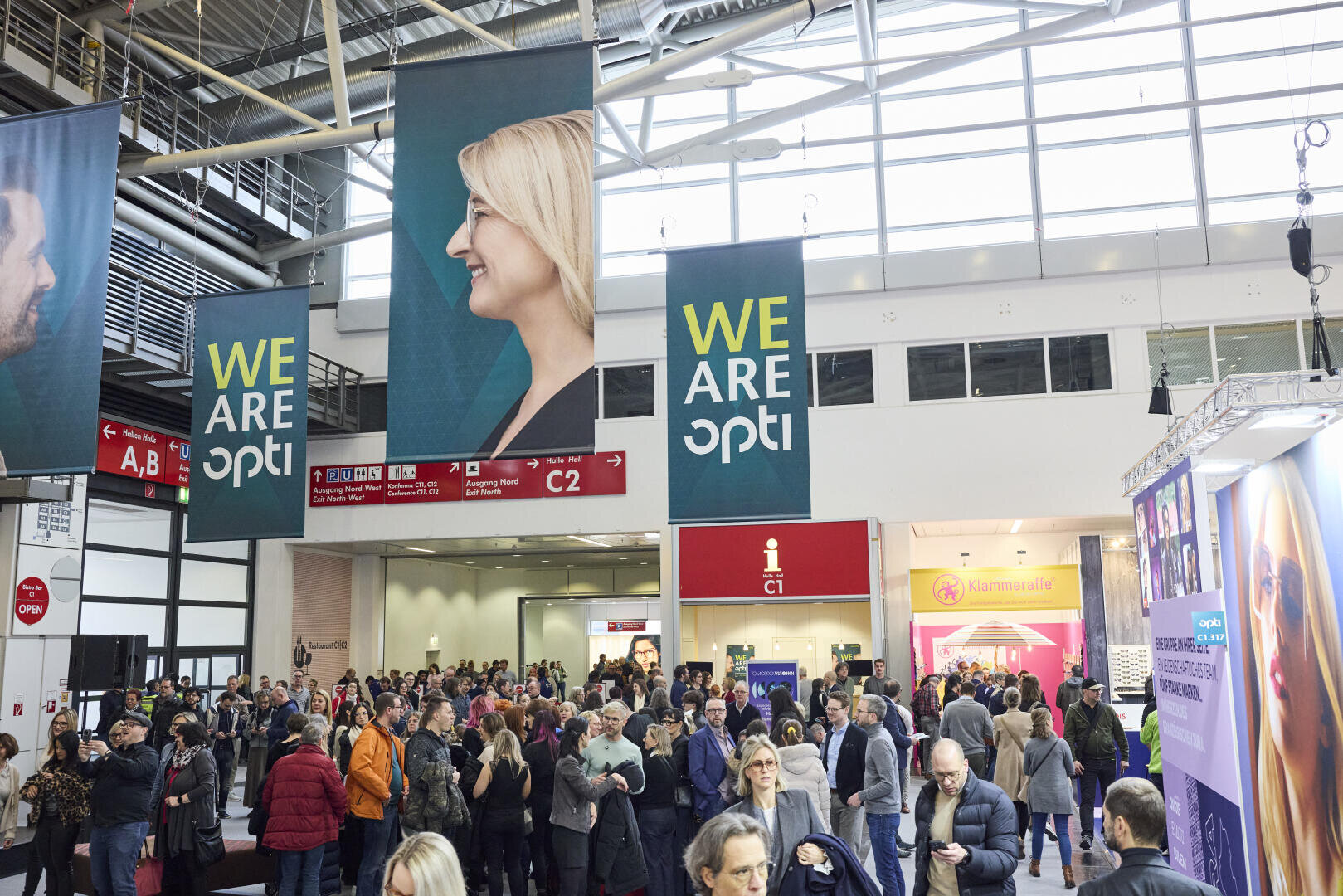 A large crowd gathers in a brightly lit convention center with WE ARE OPTI banners and smiling faces. The space has high ceilings, large windows, and directional signs hanging above the crowd.