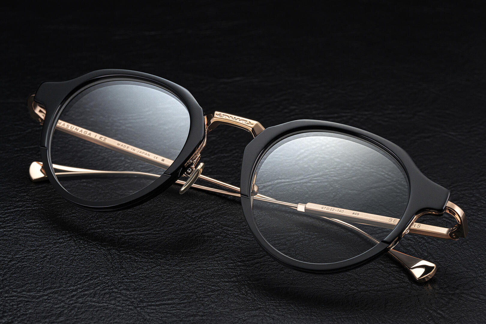 Eine stilvolle Brille mit runden schwarzen Rahmen und goldenen Metallbügeln auf einer strukturierten schwarzen Lederoberfläche.
