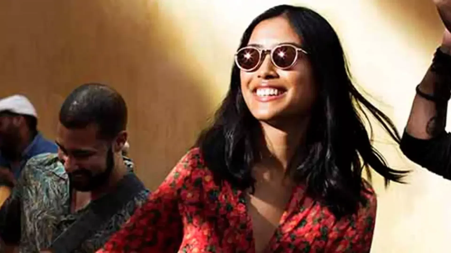 A woman with long black hair wearing sunglasses and a red floral dress smiles brightly in sunlight. A man with short hair and a beard is in the background, looking down.