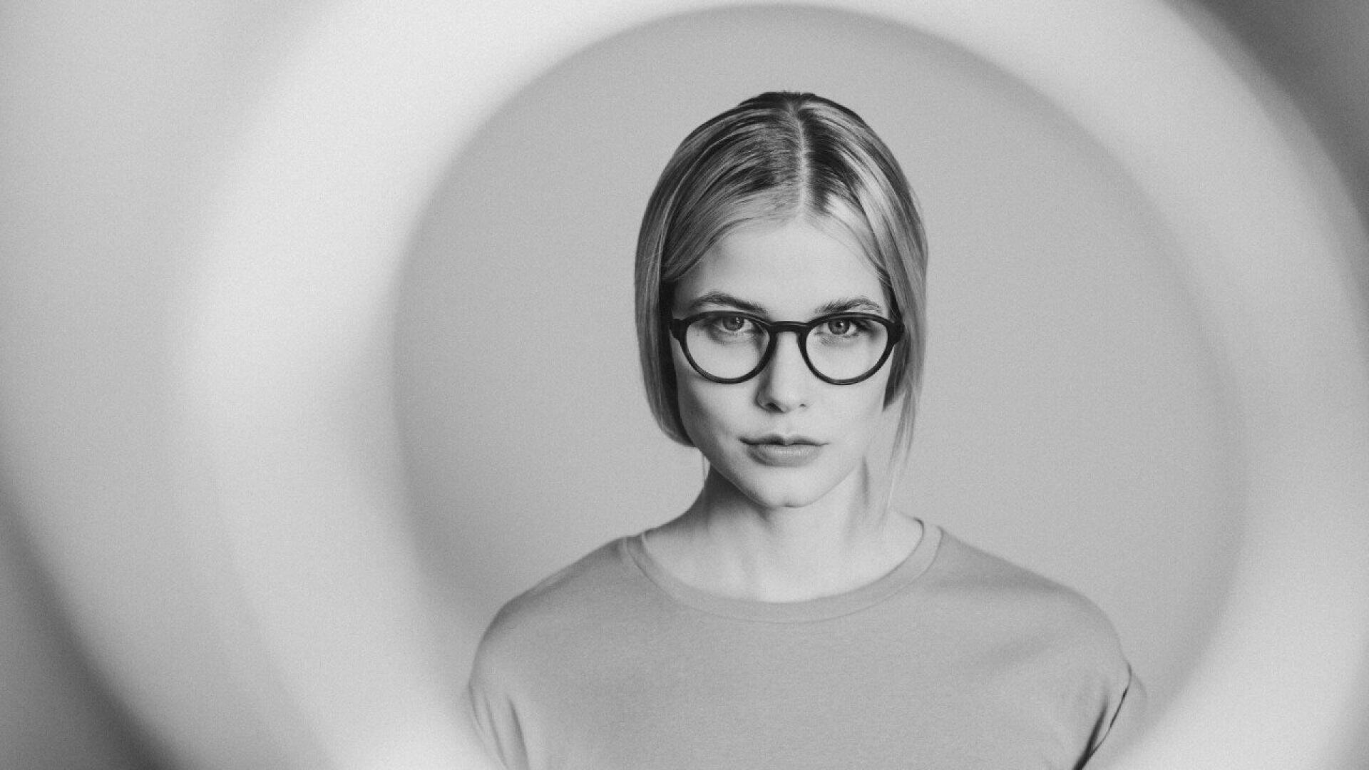 A woman with short blonde hair and round glasses looks directly at the camera, framed by a circular light in a grayscale image. She wears a plain shirt and has a neutral expression.