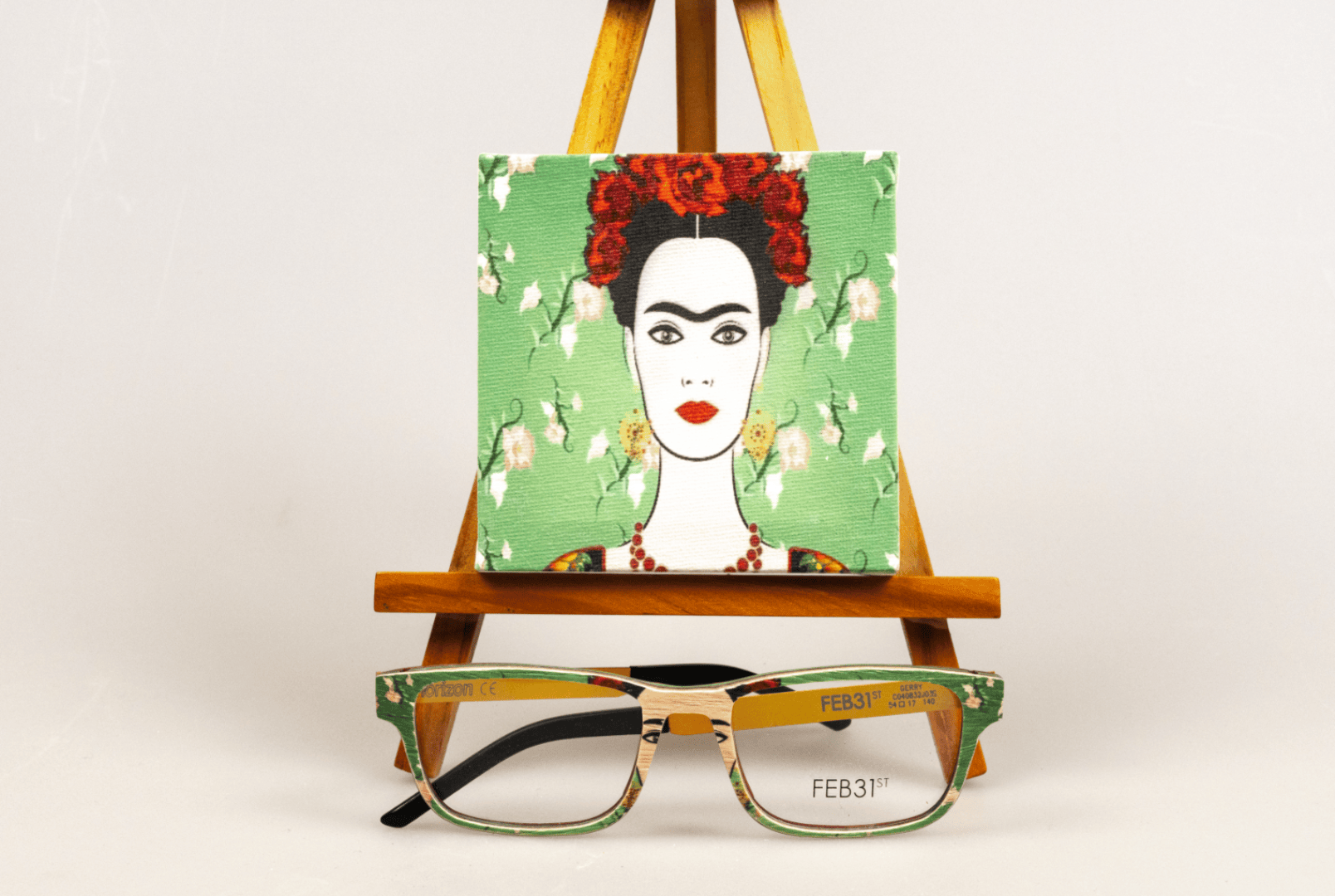 Auf einer hölzernen Staffelei steht ein kleines Leinwandgemälde von Frida Kahlo mit grünem Blumenhintergrund. Vor der Staffelei steht eine Brille mit grün gemustertem Gestell.