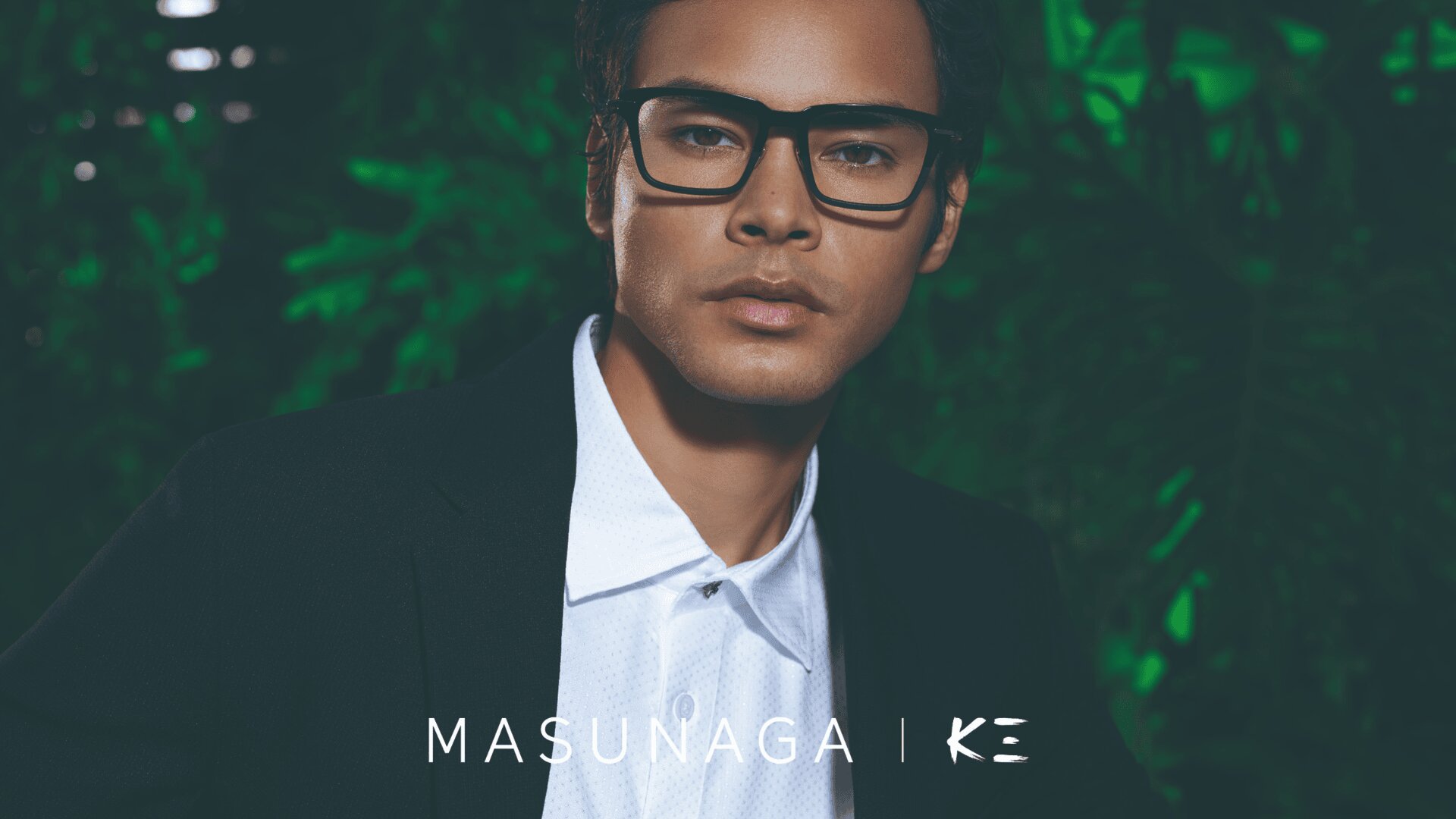 Ein Mann mit kurzen dunklen Haaren trägt eine schwarze rechteckige Brille, ein weißes Hemd und eine schwarze Jacke und posiert vor grünem Blattwerk. Der Text MASUNAGA | KE wird unten angezeigt.