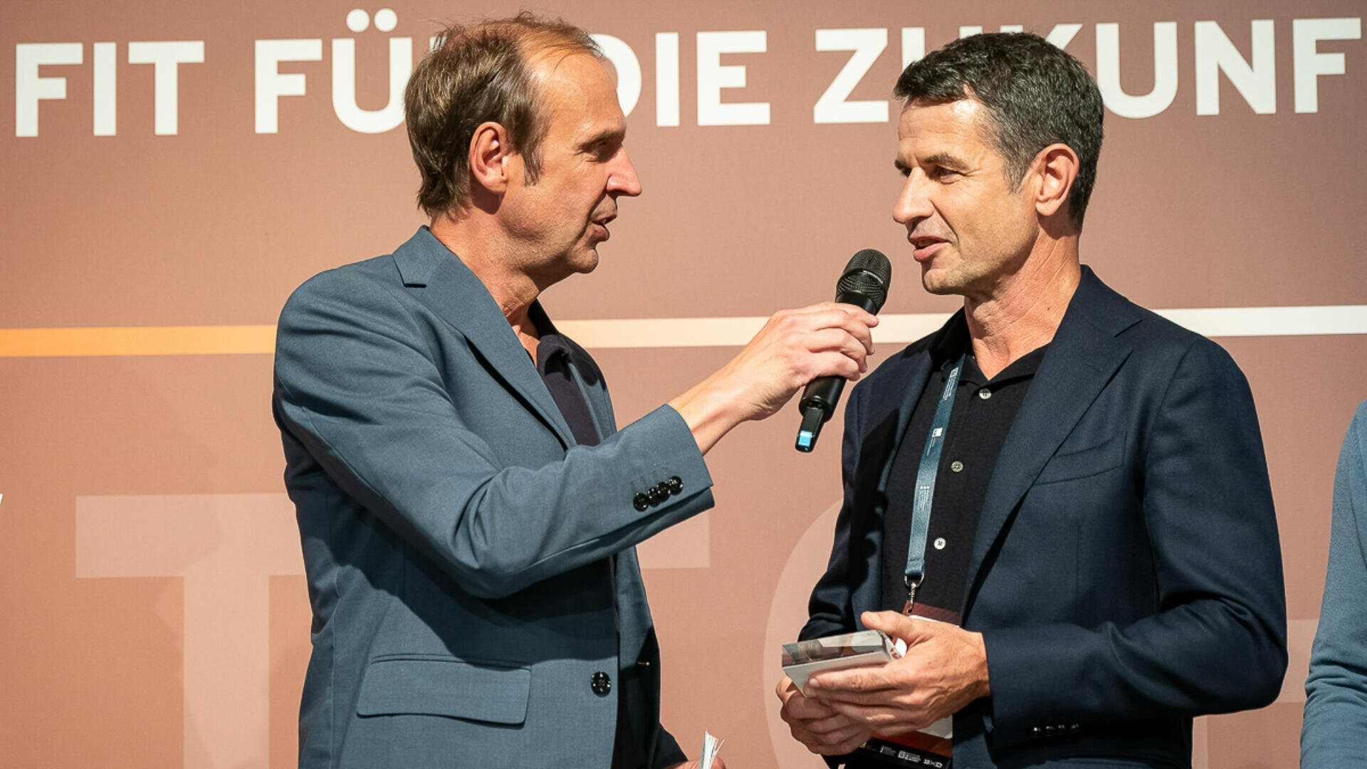 Zwei Männer in Anzügen stehen auf der Bühne. Einer hält ein Mikrofon und interviewt den anderen, der eine kleine Auszeichnung in der Hand hält. Im Hintergrund ist großer deutscher Text zu sehen. Beide wirken in ein Gespräch vertieft.