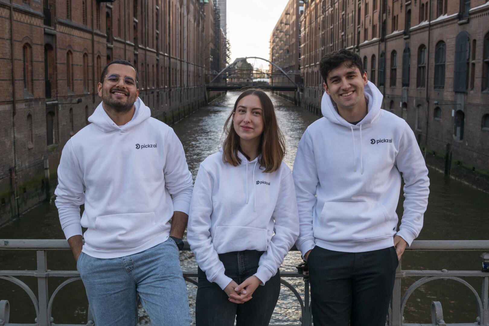 The three founders of pickz si wearing white hoodies with logos stand on a bridge in Hamburg over a canal with brick buildings on either side. They smile and pose for the photo.