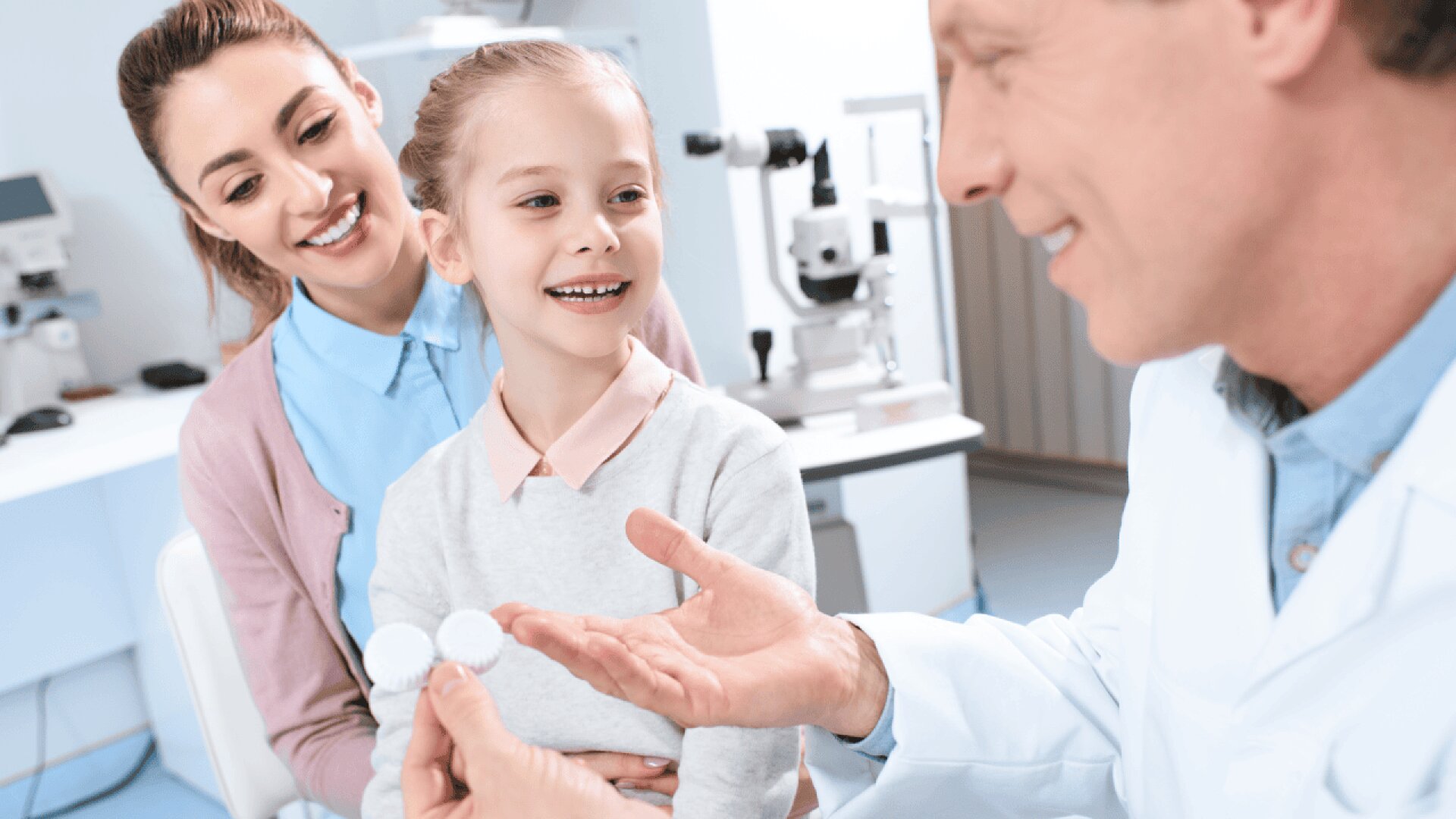 A smiling optometrist shows a contact lens case to a young girl and her smiling mother during an eye exam in a bright medical office.