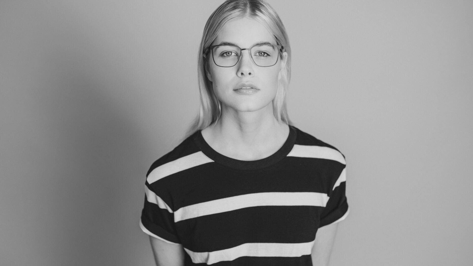 Schwarzweißfoto einer Person mit langem, glattem Haar, Brille und gestreiftem T-Shirt, die vor einem schlichten Hintergrund steht und direkt in die Kamera blickt.